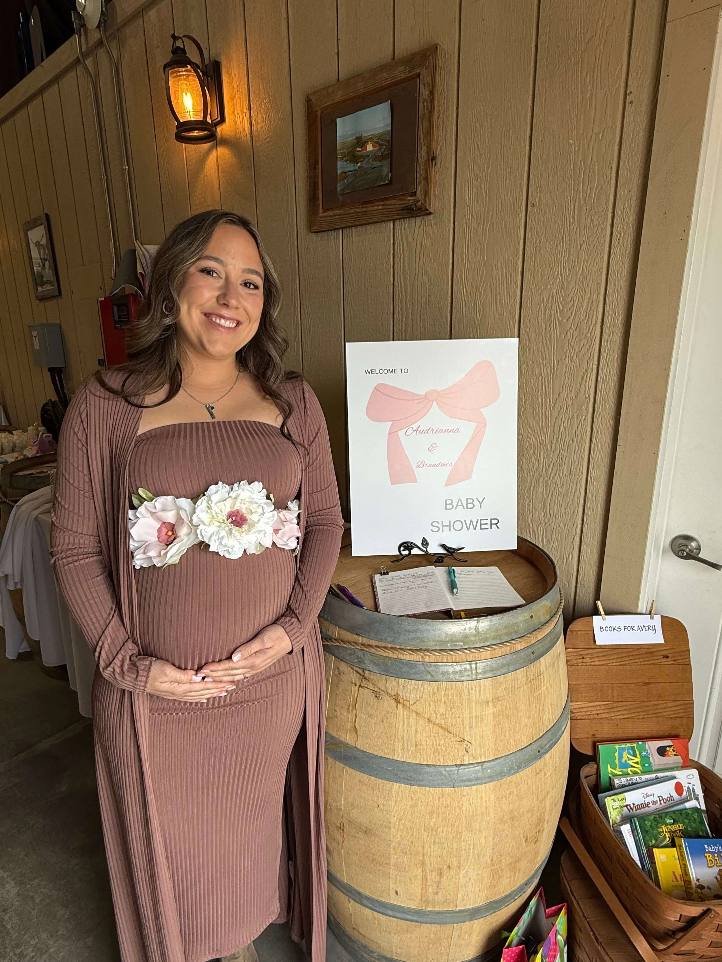 A smiling woman in a pinkish-brown dress holding a white flower bouquet, standing next to a sign that says "Welcome to Baby Shower" with a pink bow illustration, in a rustic setting with wooden walls and books for a baby shower event.