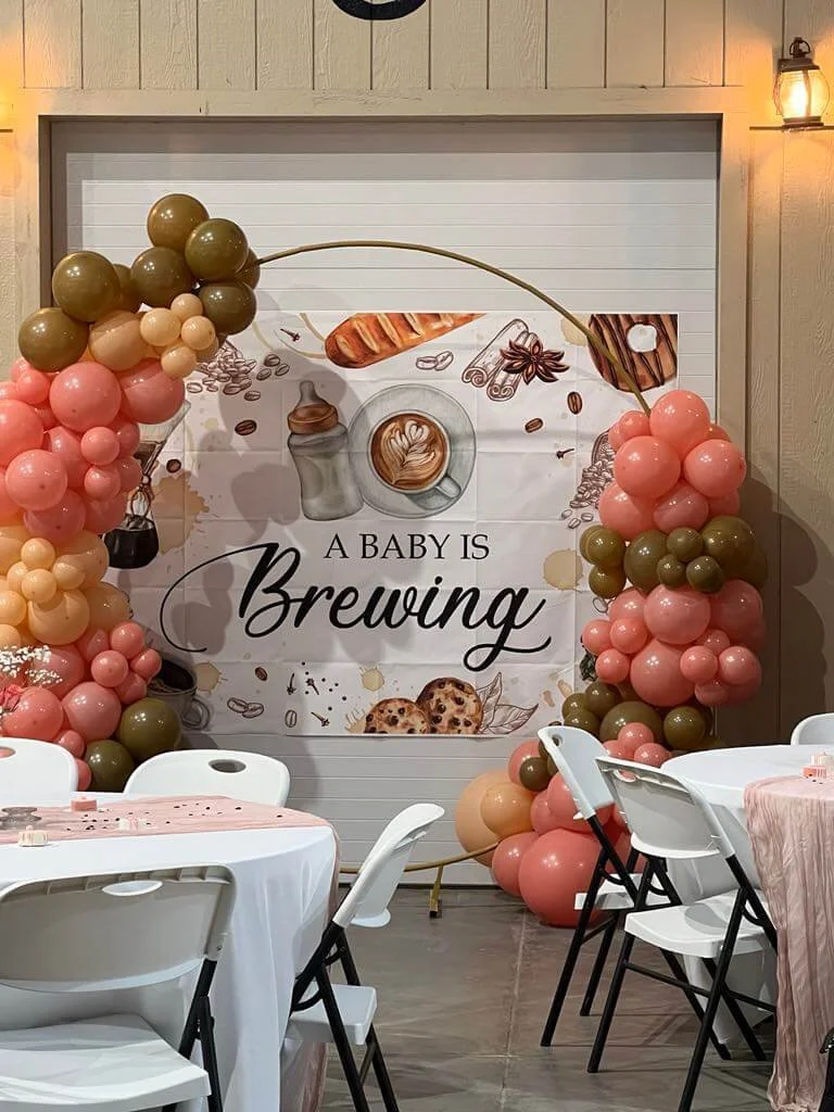 Decorative backdrop with balloon arch in pink, peach, and olive green colors, featuring a banner with the message "A BABY IS Brewing" and coffee-themed illustrations, at a baby shower event.