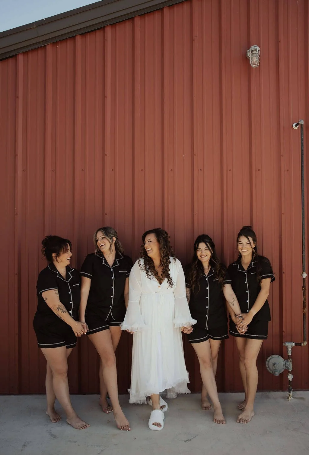 Women standing and laughing together outside in front of a red wall, dressed in matching black pajamas, with the woman in the center wearing a white dress.