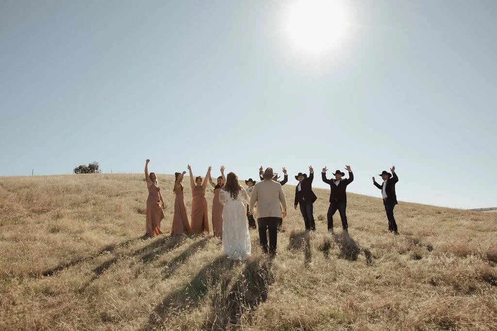 A wedding party walking and dancing on a grassy field under a clear sky with the sun shining.