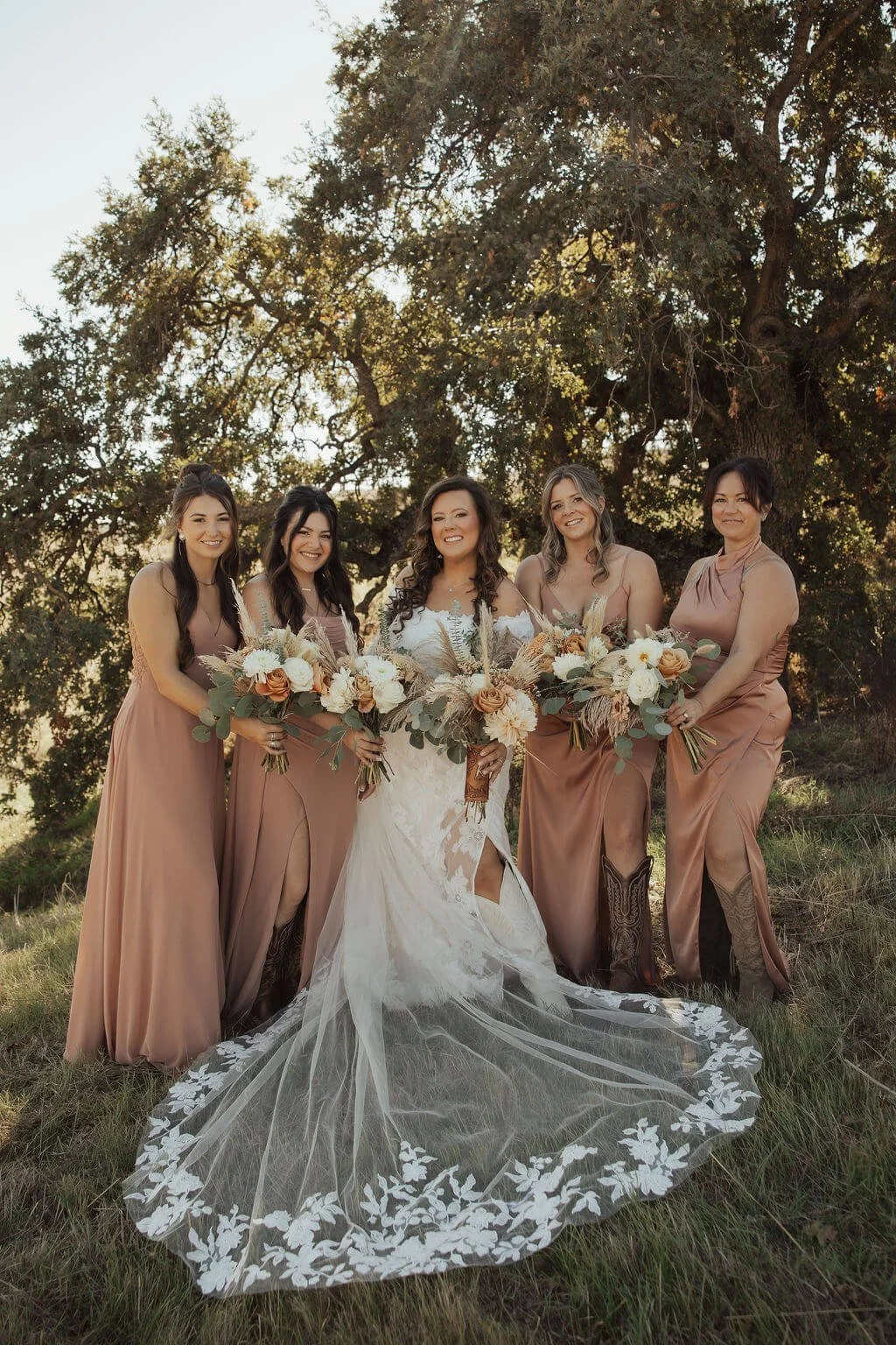 Bride and four bridesmaids standing outdoors, holding bouquets, under a large tree in sunlight.