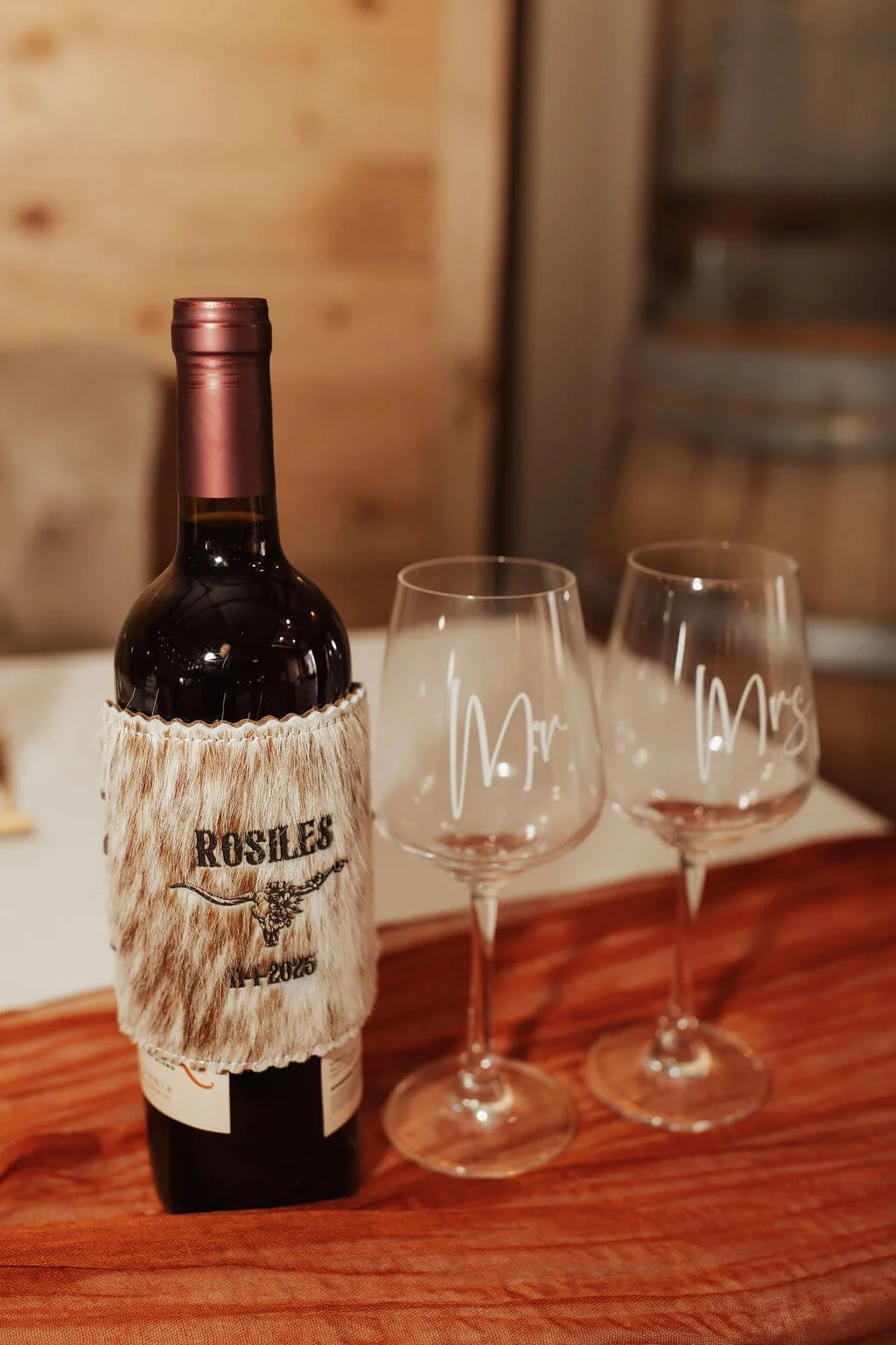 A bottle of red wine with a fell fabric label that reads "Rousles" and two empty wine glasses with white script writing, one marked "Mr" and the other "Mrs". They are on a wooden surface with a blurred wooden wall background.