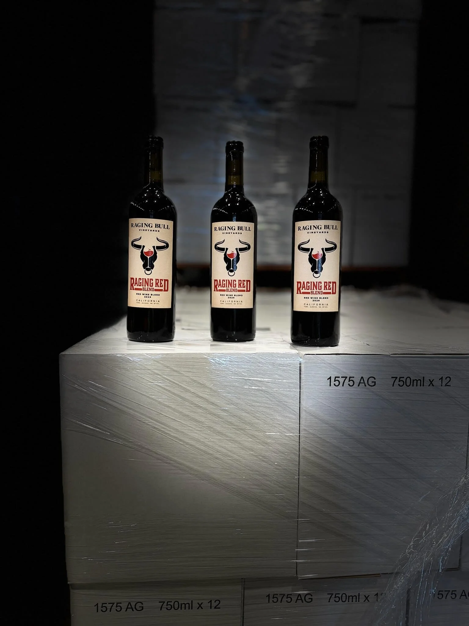 Three bottles of Raging Bull Vineyards Raging Red Blend wine on a white surface. The background is dark, with a textured wall.