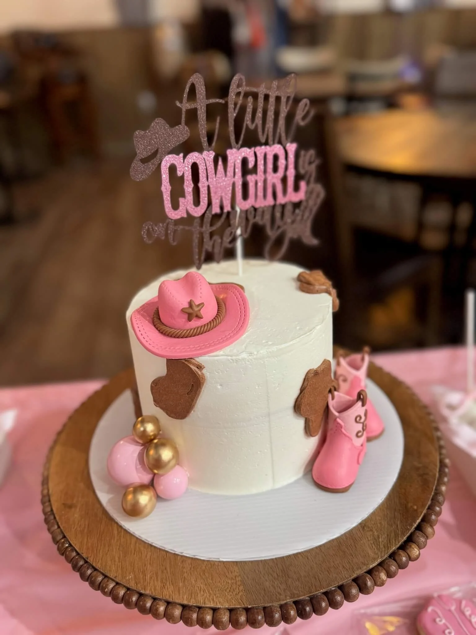 A white cake decorated with pink and gold cowboy-themed decorations, including a pink cowboy hat, pink cowboy boots, and gold and pink balls, with a topper that reads 'A Little Cowgirl' in glittery pink and brown lettering.