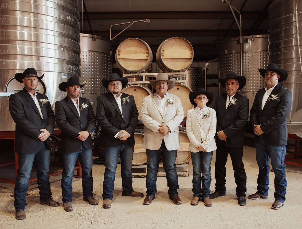 A group of seven men and one boy dressed in western attire, including cowboy hats, suits, and badges, standing in front of large stainless steel fermentation tanks and wooden wine barrels inside a winery or brewery.