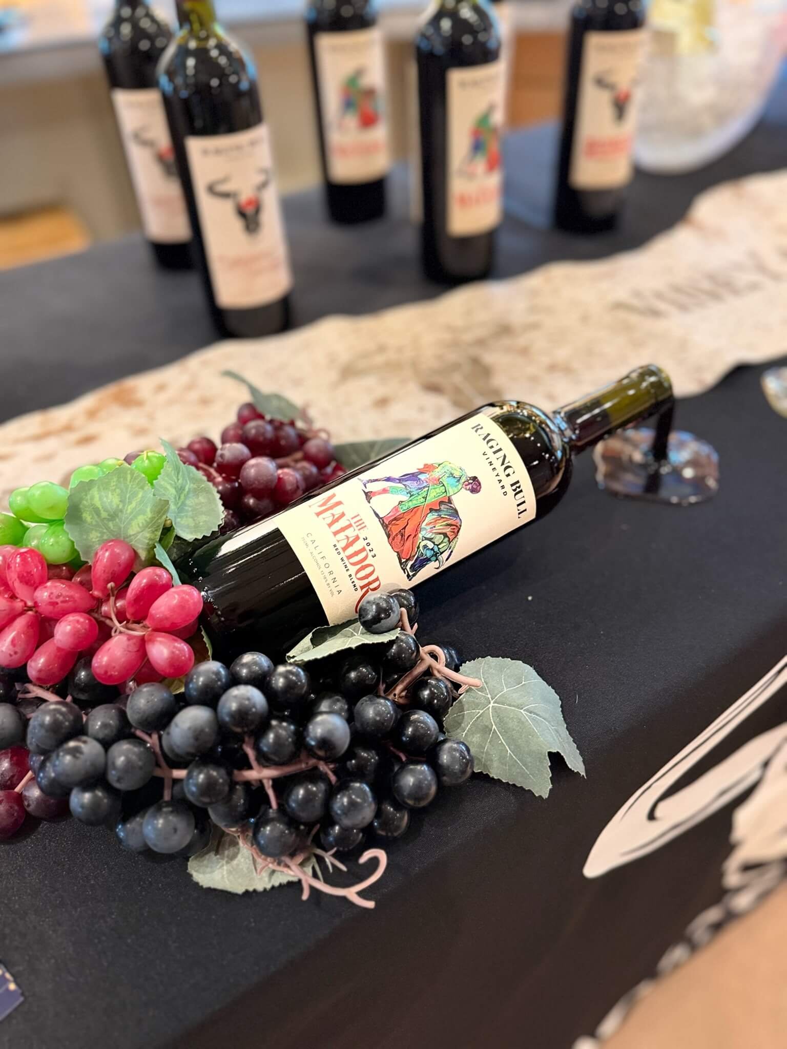 A display of a red wine bottle lying on a table decorated with artificial grapes and leaves, with blurred bottles in the background.