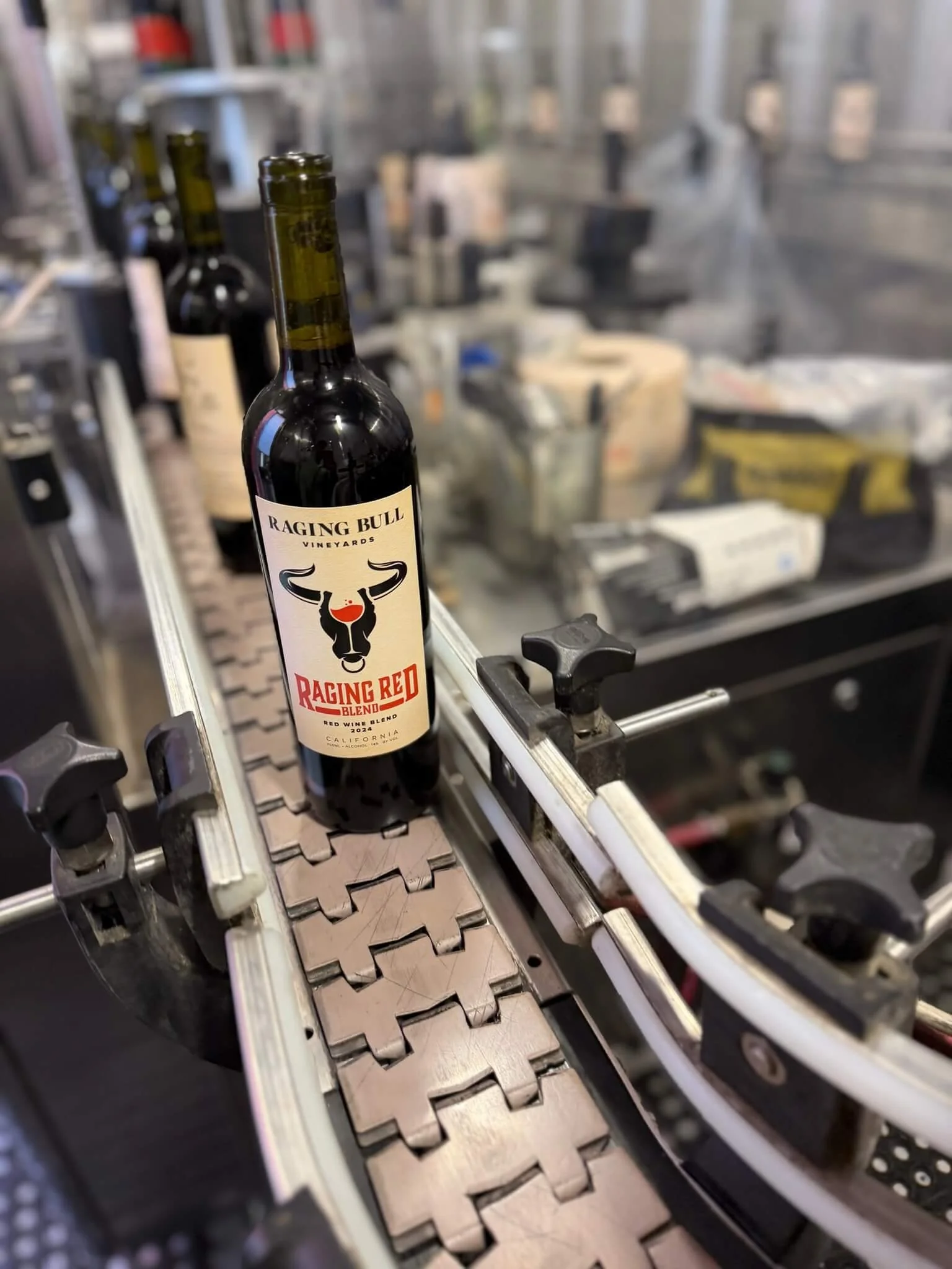 A wine bottle labeled 'Raging Bull Vineyards Raging Red Blend' appears on a conveyor belt in a winery or bottling facility, with blurred industrial equipment and shelves in the background.