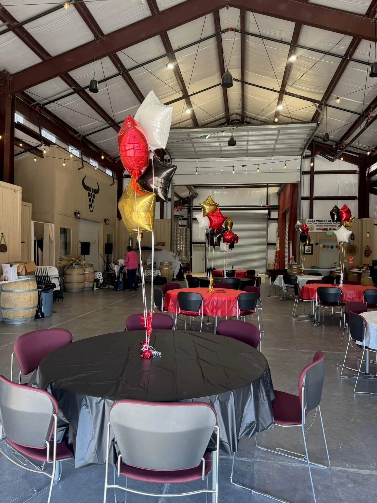 Indoor event space decorated with balloons in red, black, white, and gold, and tables covered with black and red tablecloths for a celebration.