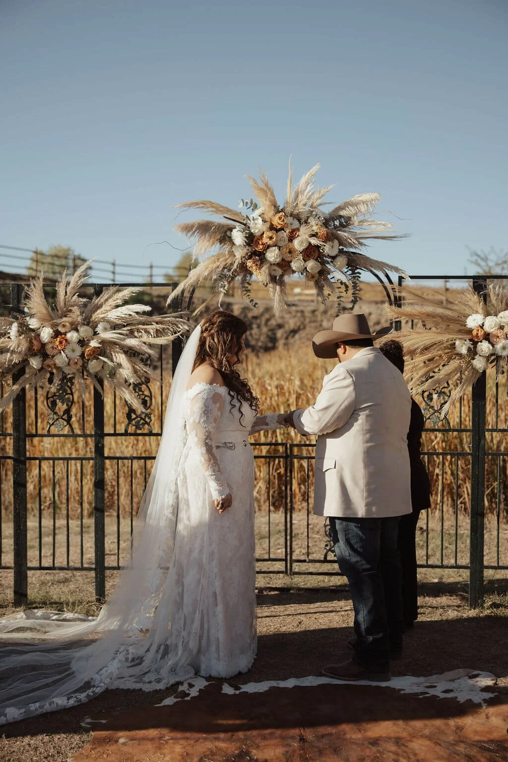 A wedding ceremony outdoors with a bride and groom exchanging vows under a decorated arch with pampas grass and flowers, surrounded by a rural landscape and a clear blue sky.