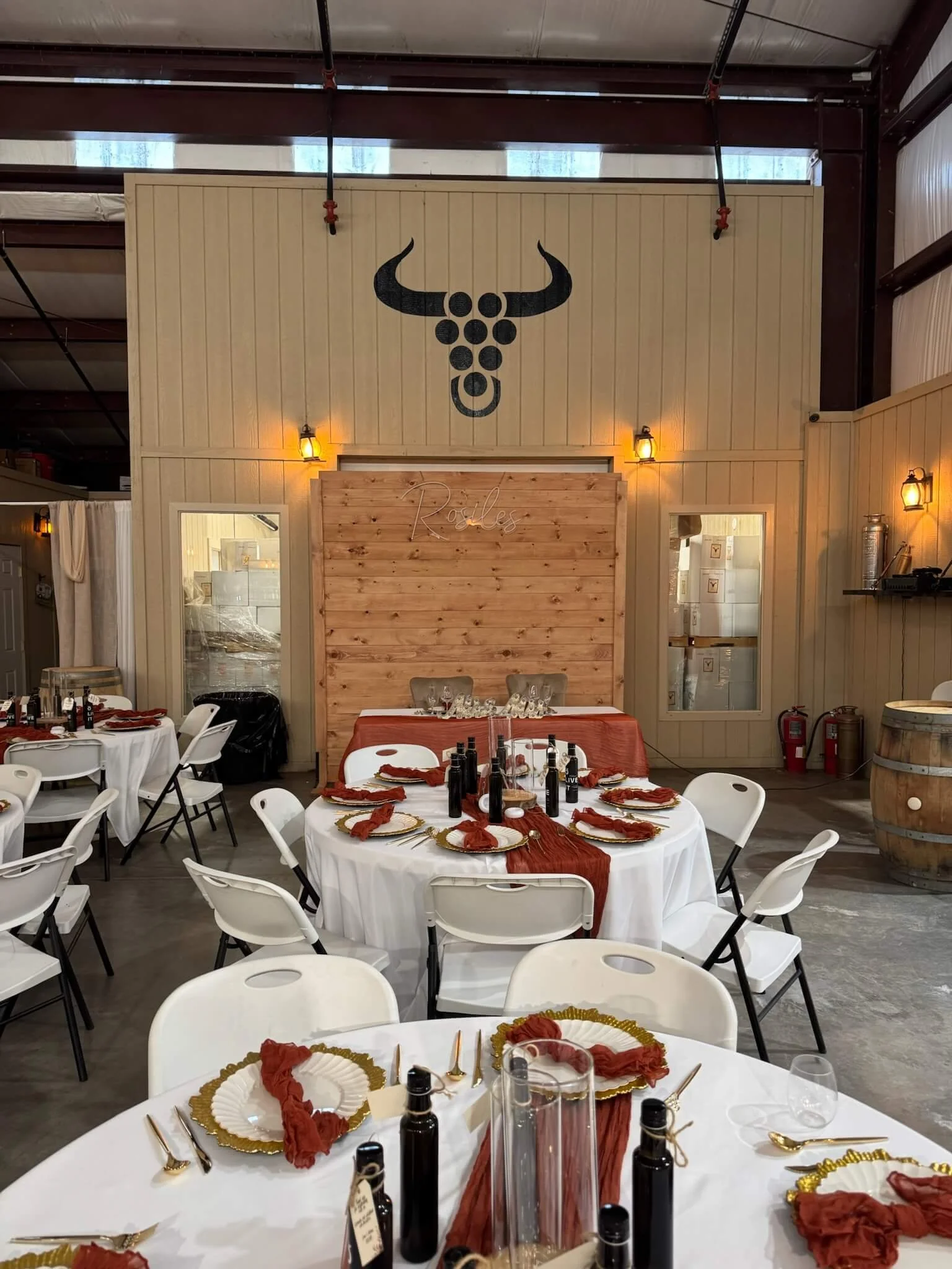Indoor wedding reception setup with round tables covered in white tablecloths, decorated with red table runners, gold charger plates, and red napkins. Wine bottles and glasses are arranged on the tables. Wooden wall with two windows and a sign reading 'Rules' in cursive, with string lights and wall-mounted lamps providing warm lighting. The ceiling features visible metal beams and vents.