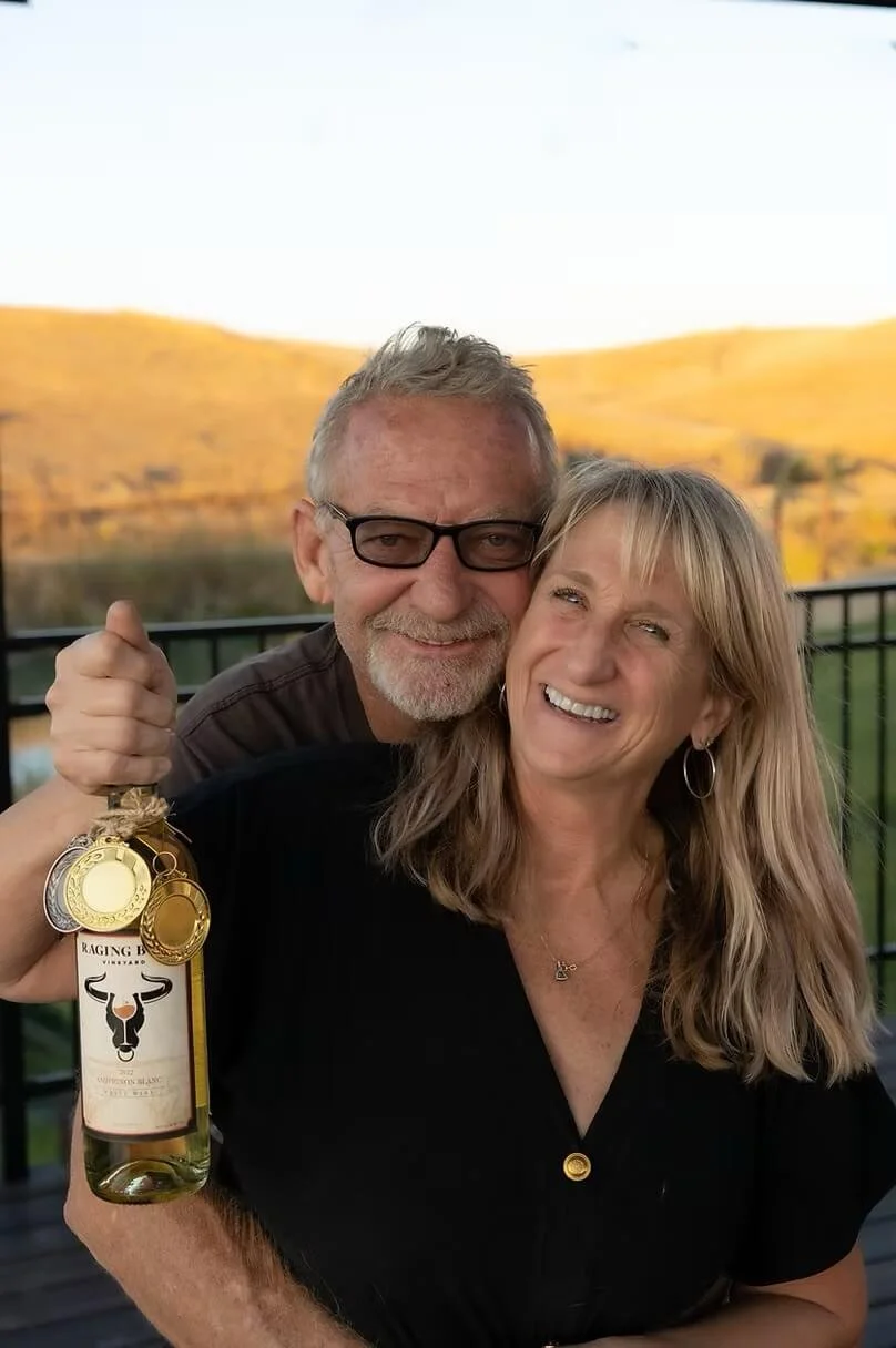 A smiling middle-aged man and woman stand outdoors on a balcony or patio, with a scenic, hilly landscape in the background. The man holds a bottle of wine.