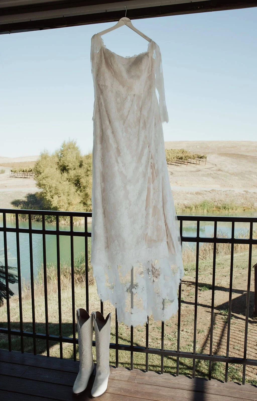 A white lace wedding dress hanging on a hanger outdoors, with a pair of white cowboy boots placed below it on a wooden deck, overlooking a landscape with trees, water, and hills in the background.