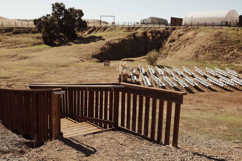 Outdoor scene with carpeted stairs leading down to a grassy area with rows of white lawn chairs arranged for an outdoor event, a rocky hillside in the background, and a greenhouse structure on the right.