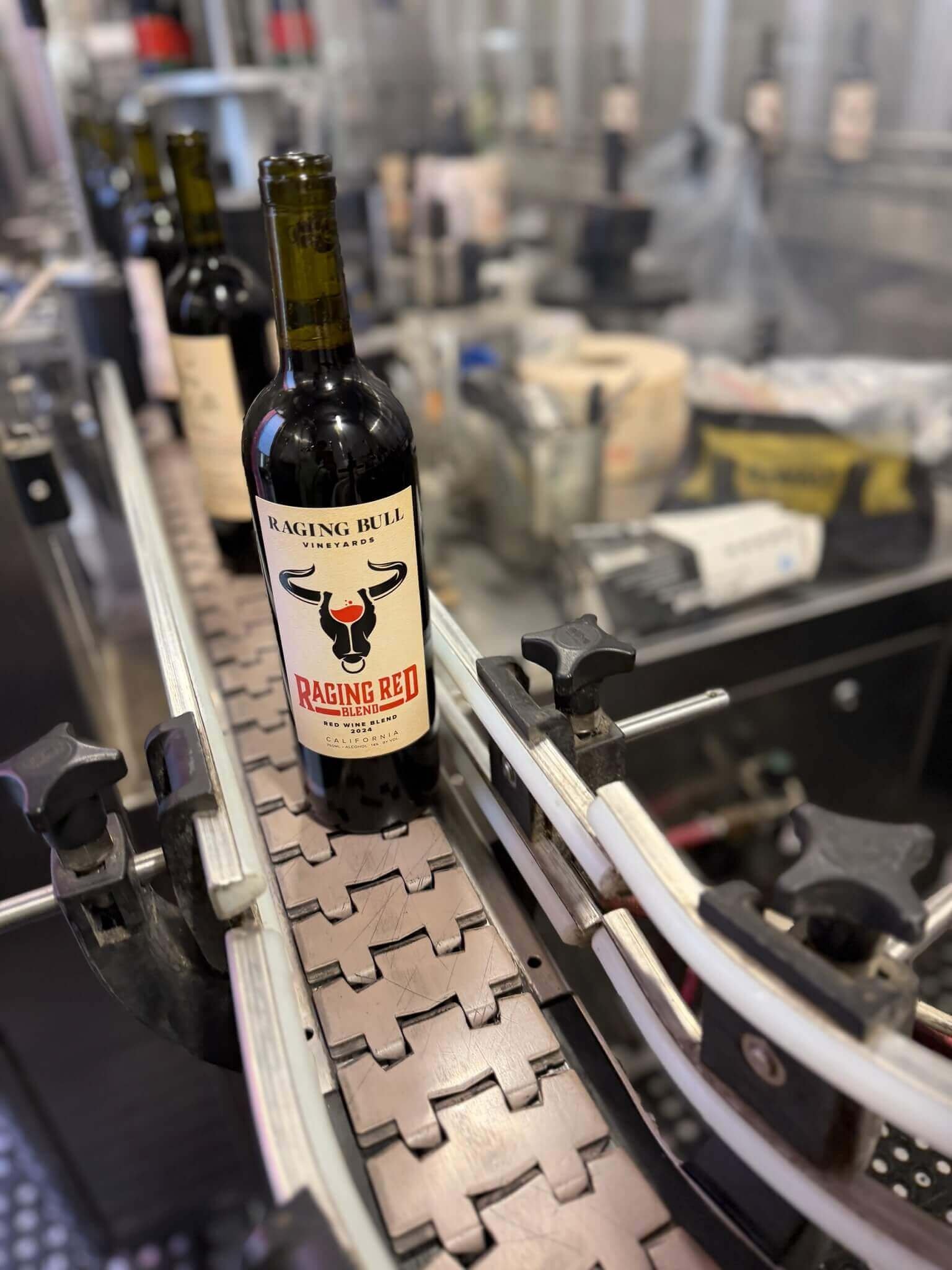 A wine bottle labeled 'Raging Bull Vineyards Raging Red Blend' appears on a conveyor belt in a winery or bottling facility, with blurred industrial equipment and shelves in the background.
