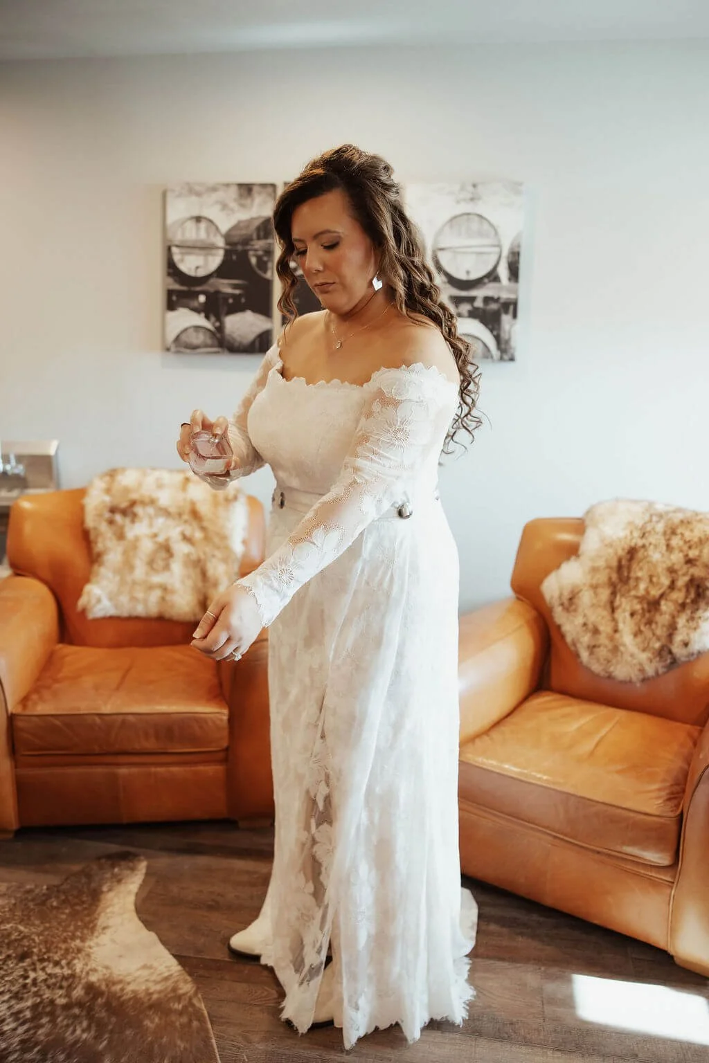 A woman in a white lace wedding dress standing in a living room, holding a small item in her hand, with leather sofas and decorative pillows behind her.