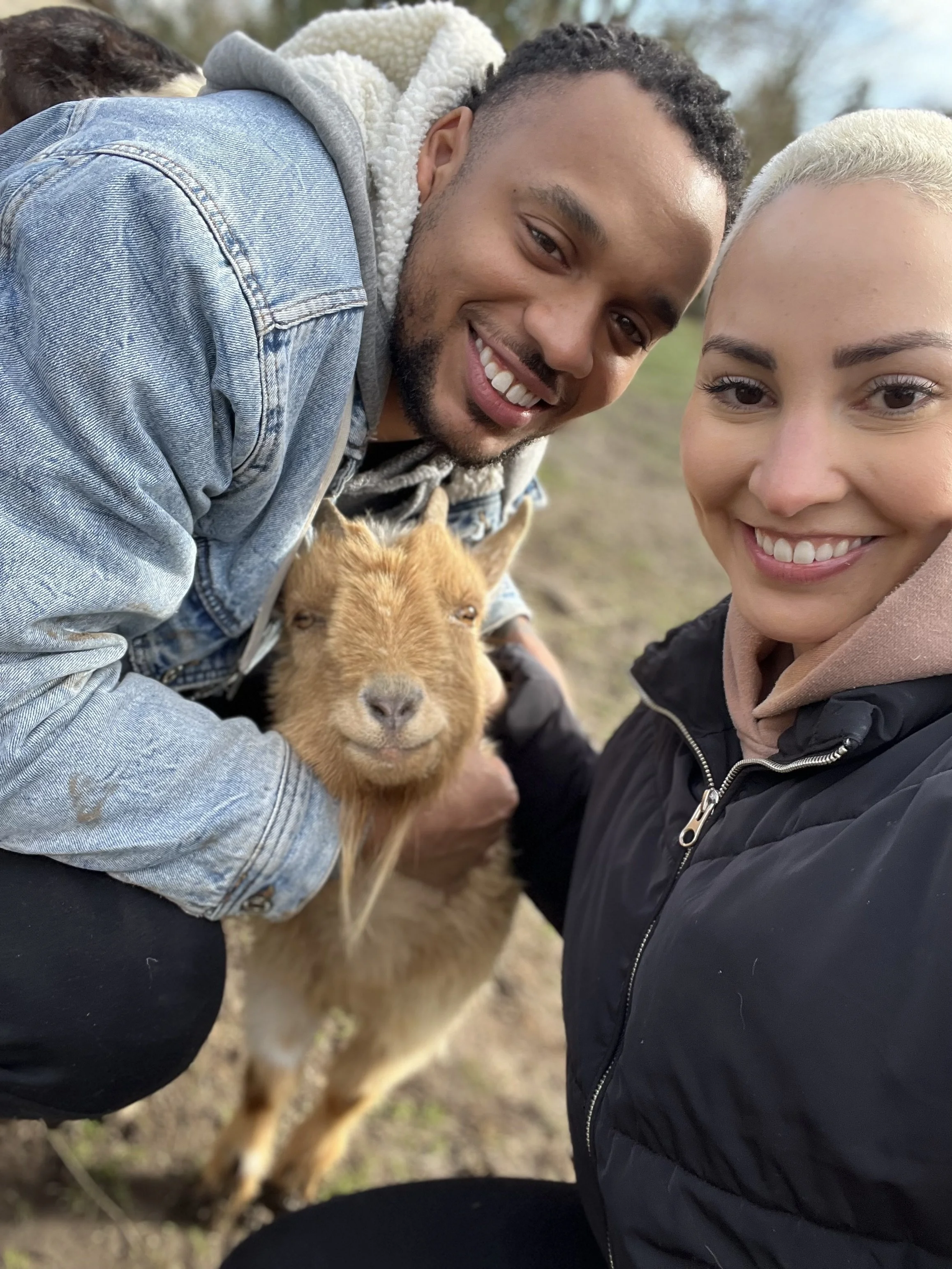 A smiling man with short curly hair and a beard and a smiling woman with blonde hair and a black jacket are taking a selfie outdoors with a happy dog between them. The dog has light brown fur and closed eyes.