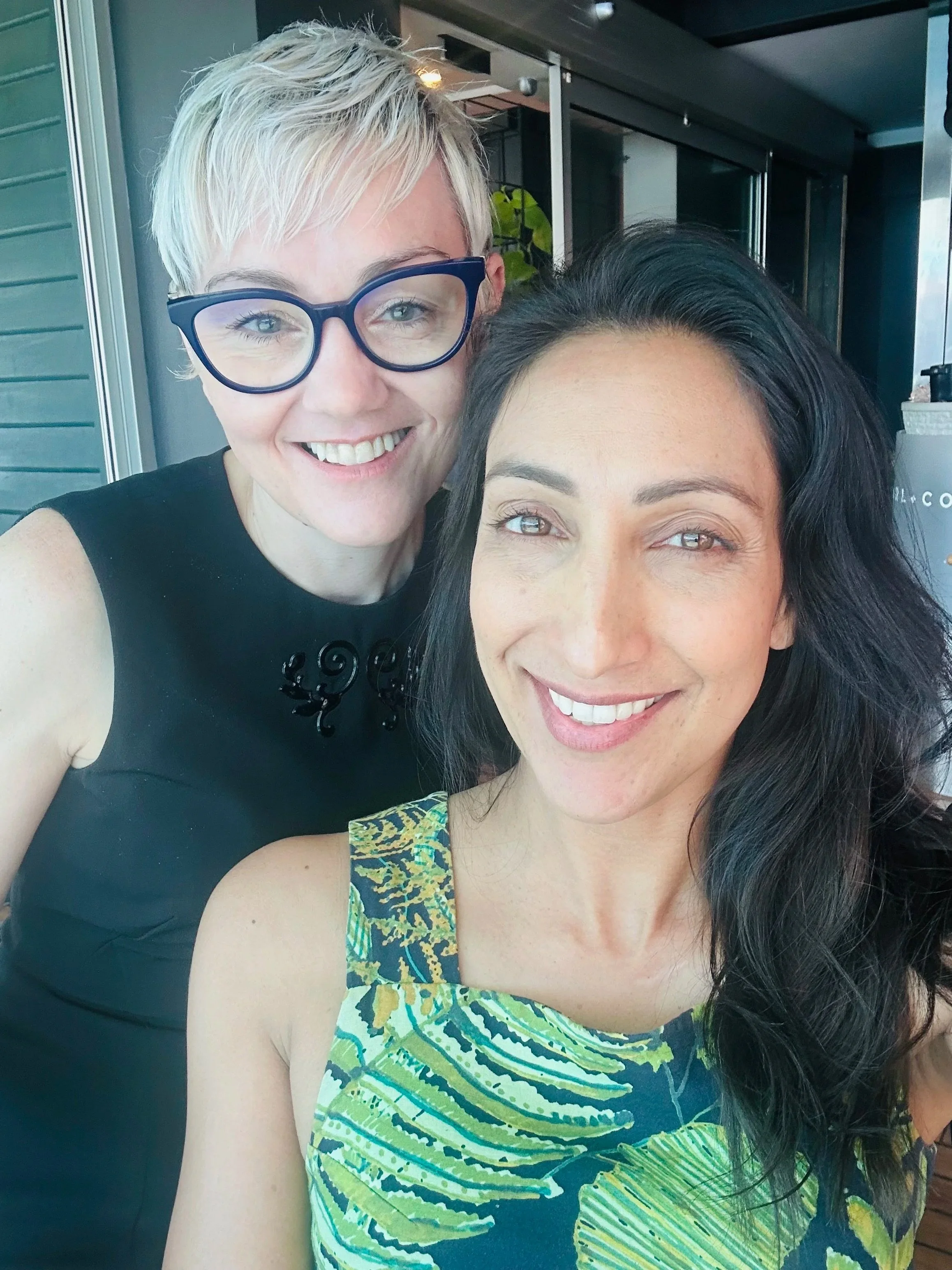 Close-up of two smiling women taking a selfie together, one with short blonde hair wearing glasses and a black top, the other with long dark hair wearing a colorful patterned dress.