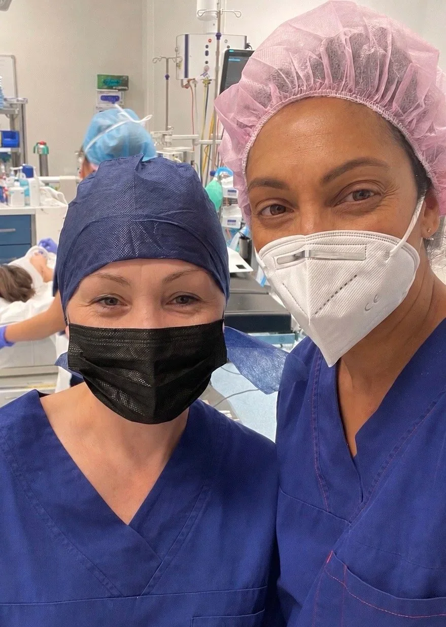 Two healthcare professionals in surgical scrubs and masks taking a selfie in an operating room with medical equipment and a patient in the background.