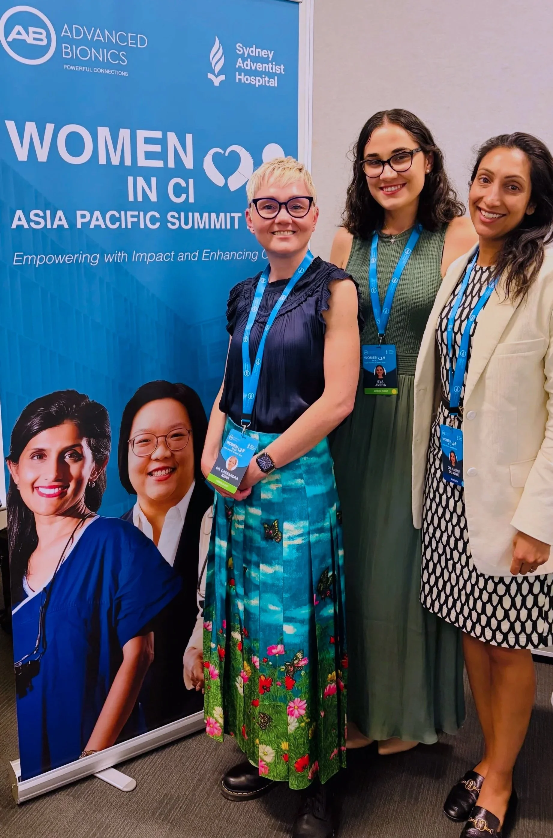 Three women standing in front of a blue banner at the Women in CI Asia Pacific Summit. The banner displays event sponsors and logos, including 'Advanced Bionics' and 'Sydney Adventist Hospital'. The women are smiling and wearing conference badges.