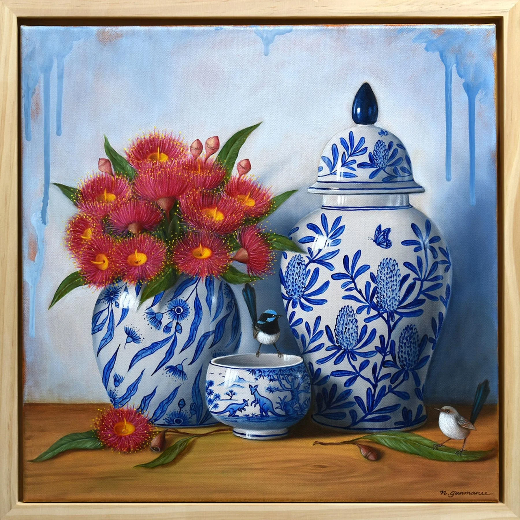 A framed oil painting of a blue and white floral-patterned vase with red gum flowers, accompanied by a small bird on a wooden table, painted by Natasha Junmanee.