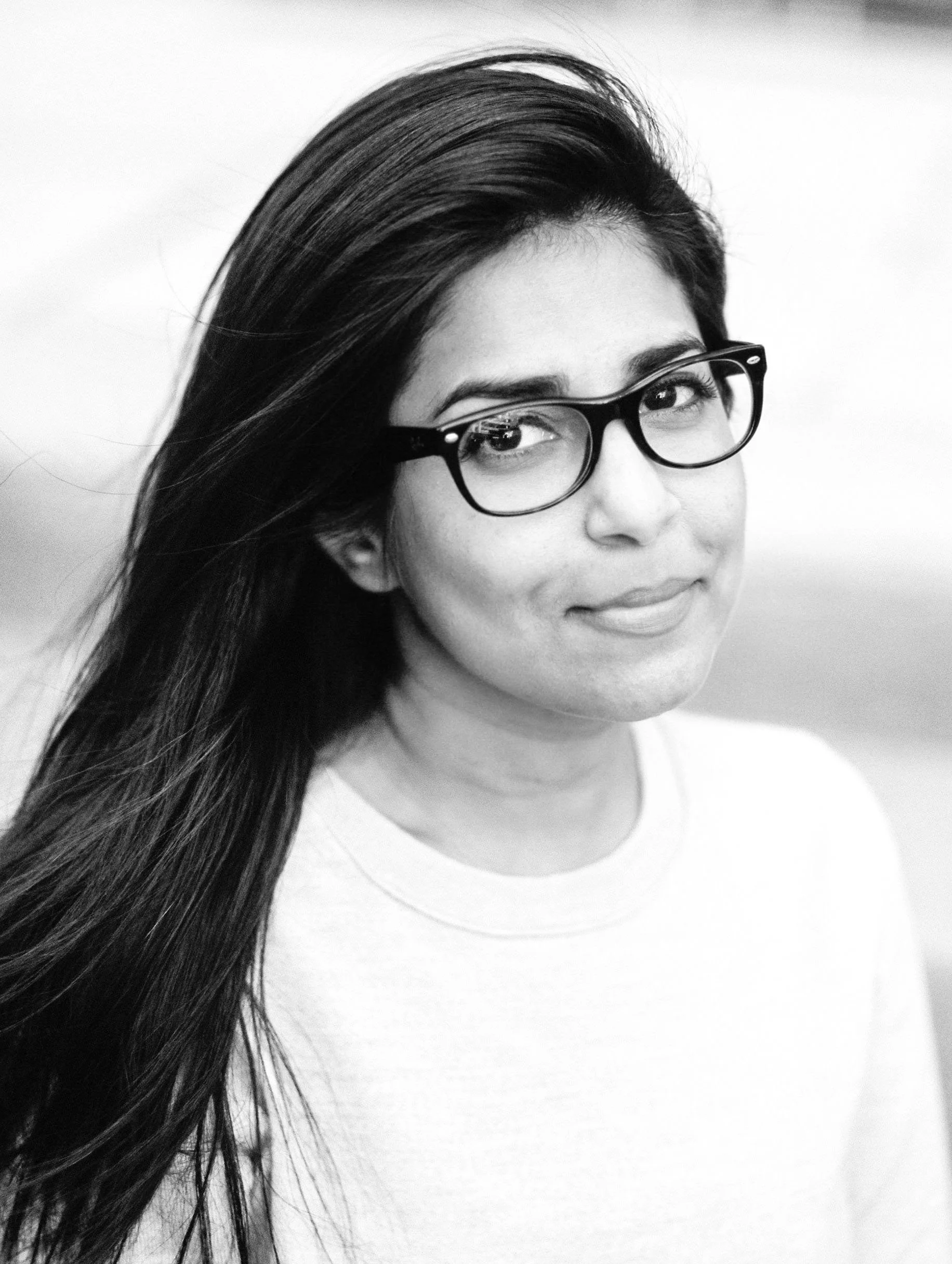 A young woman with long dark hair and wearing glasses, smiling slightly, facing the camera against a plain background.