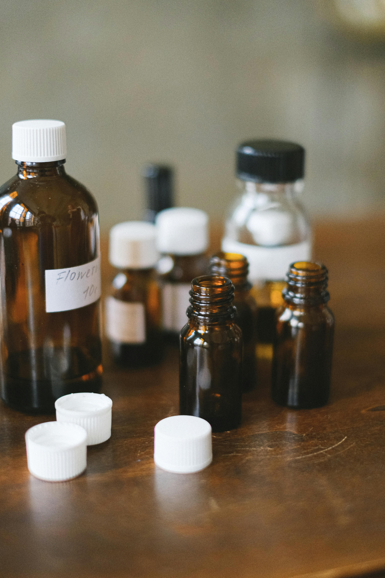 A collection of small dark brown and clear glass bottles with white caps, some labeled, arranged on a wooden surface. Some caps are off and placed in front.