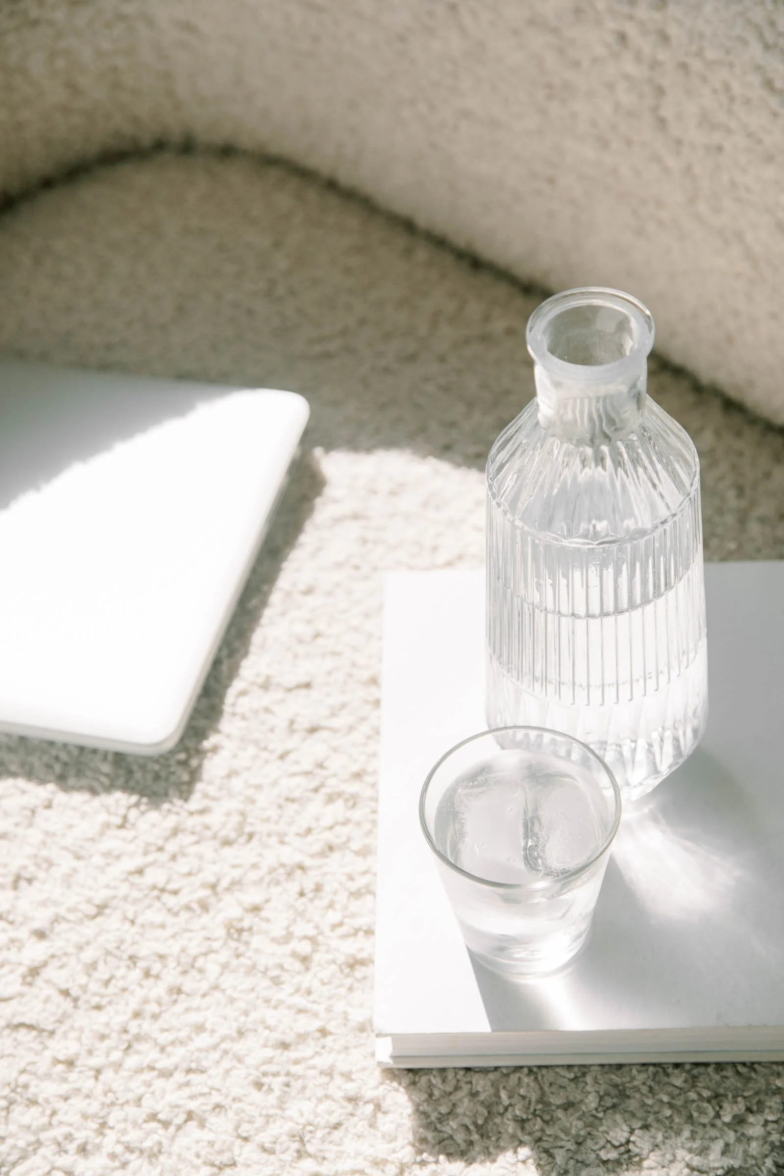 A clear glass bottle with a textured surface and a narrow neck, positioned next to a tall glass of water, on a white surface with a mirror and a beige textured wall in the background.