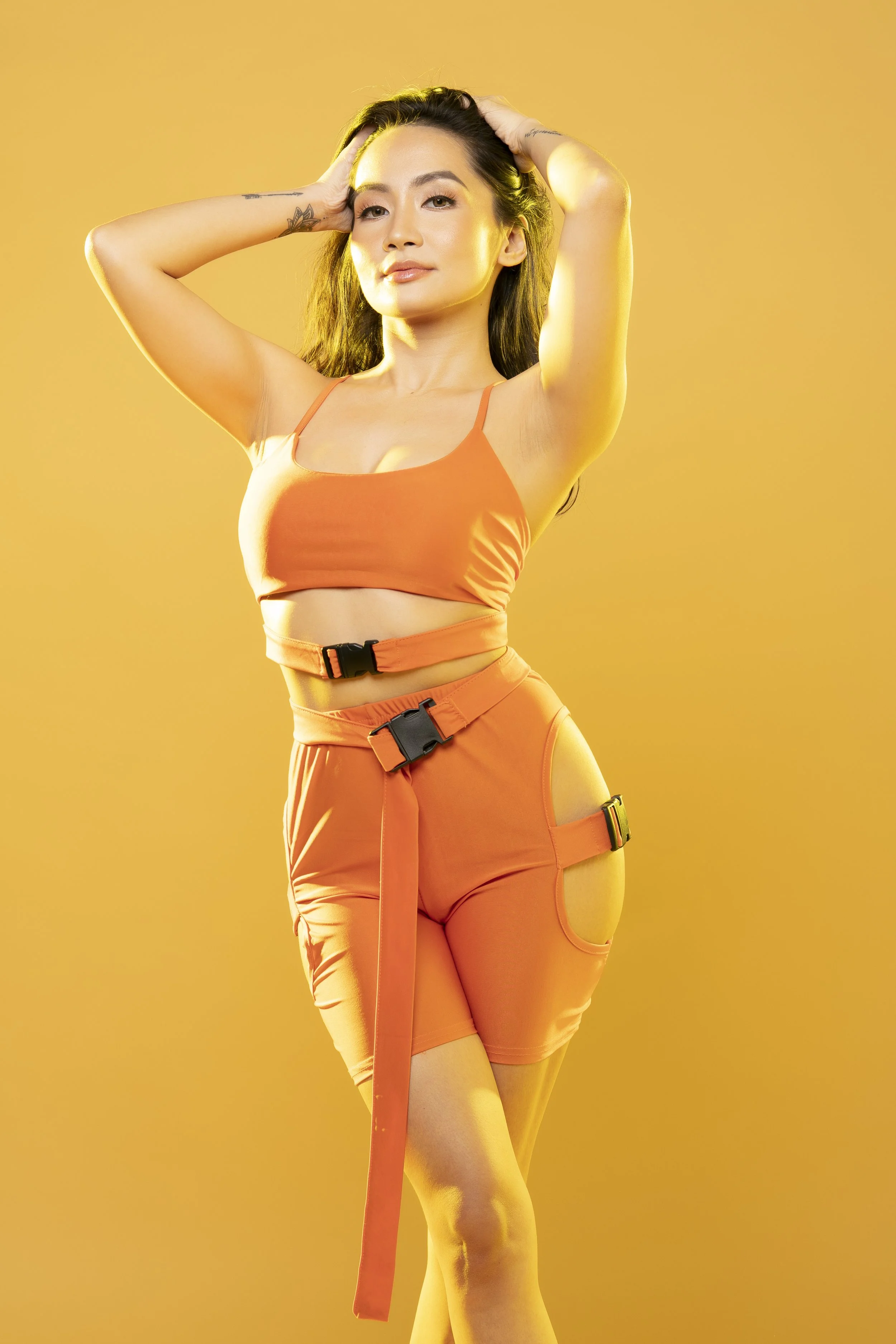 A woman with long dark hair and tattoos poses with her hands in her hair against a yellow background, wearing an orange crop top and orange pants with black buckle accents.
