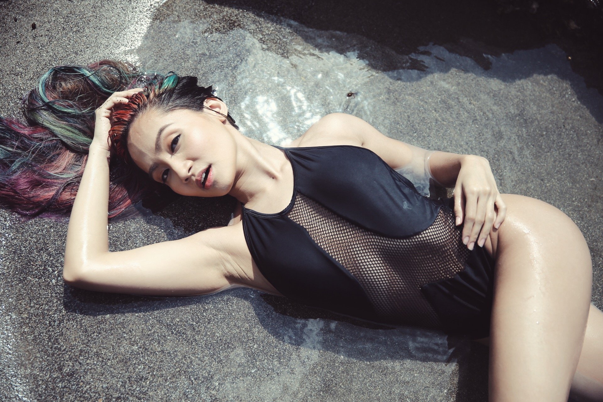 Woman lying on wet sandy beach with her head resting on her hand, wearing a black mesh swimsuit, with colorful hair and a relaxed expression.