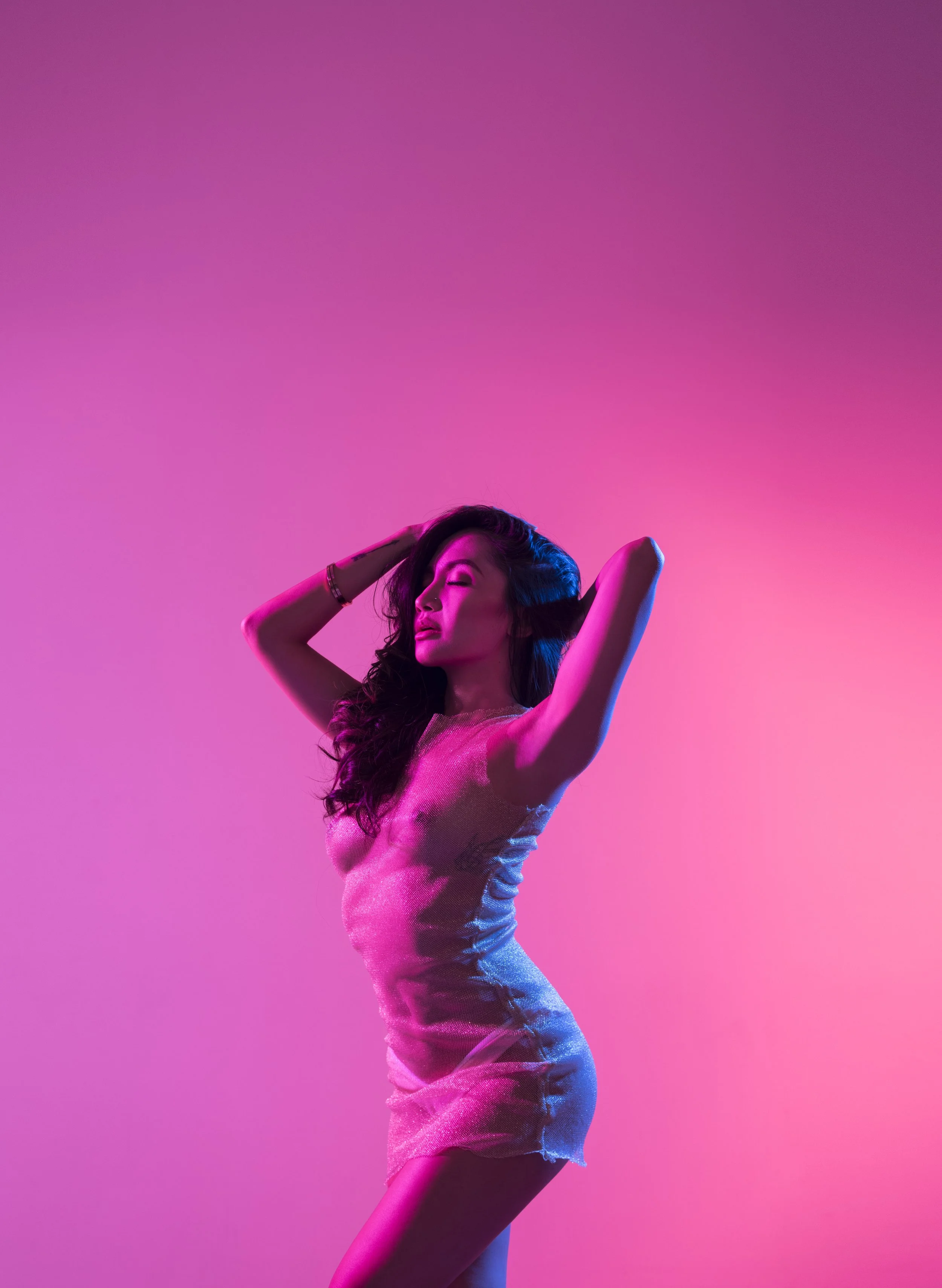 A woman with long dark hair and closed eyes posing with her hands behind her head, wearing a shiny sleeveless dress, against a gradient pink and purple background.