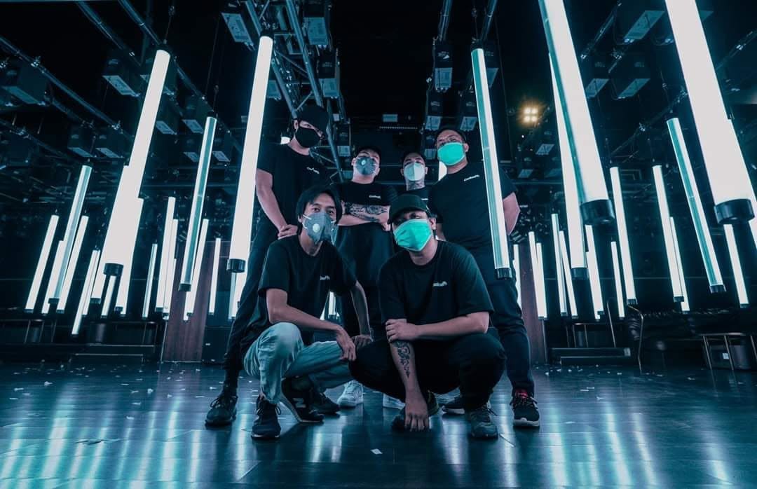 Six people wearing black shirts and masks, posing in a dark, futuristic room with vertical neon lights and reflective floor.