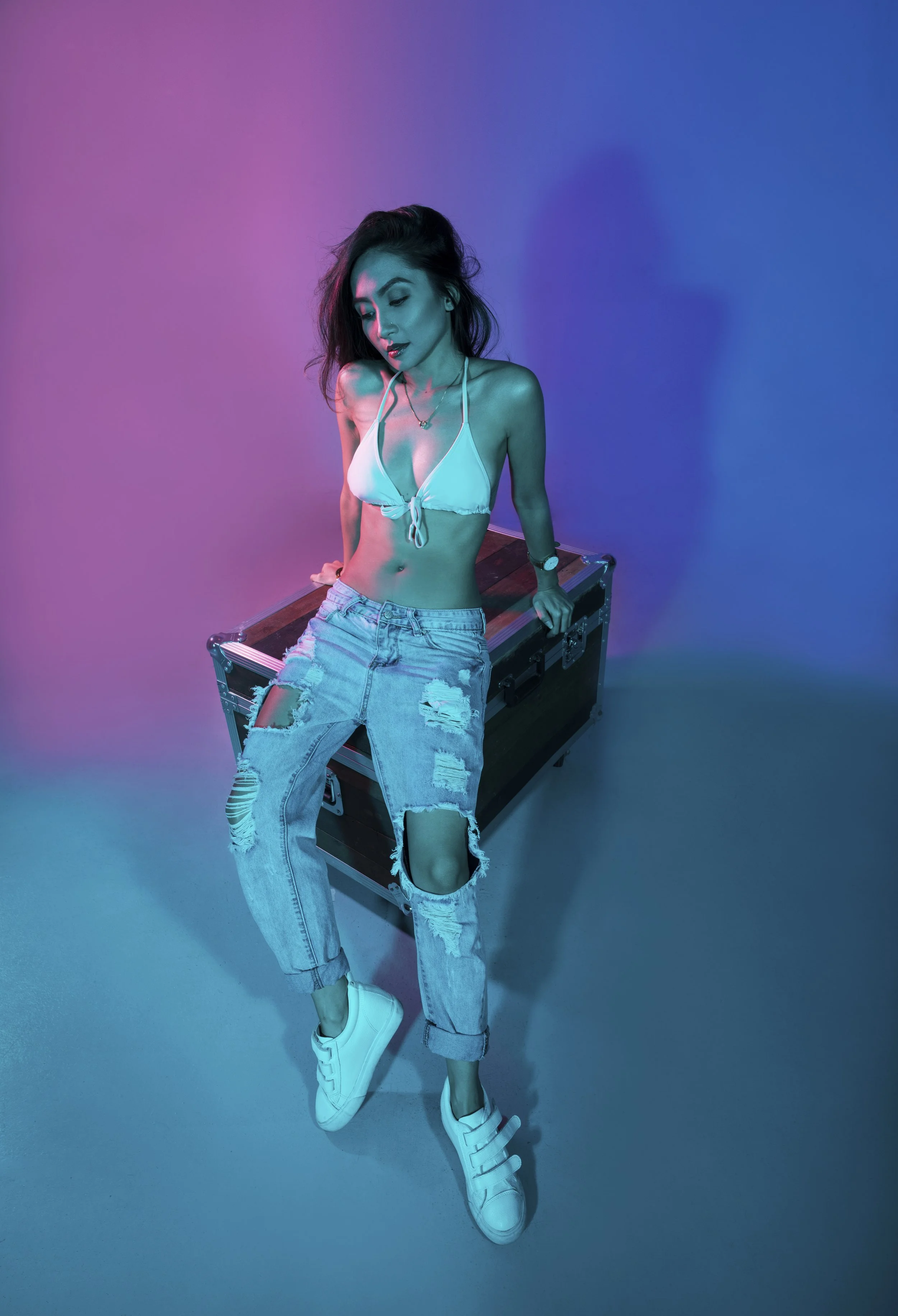 A young woman with shoulder-length hair, wearing a white bikini top, ripped jeans, and sneakers, posing with one knee bent and leaning on a black equipment case against a multicolored background.