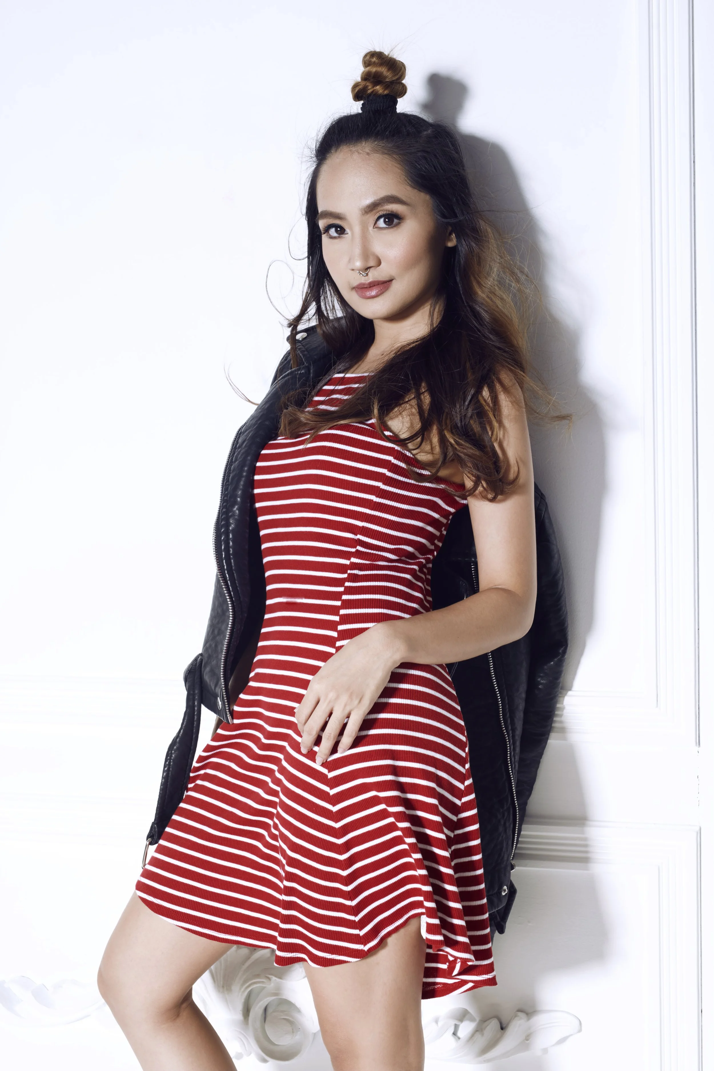 Young woman in a red and white striped dress, white sneakers, and a black leather jacket, standing against a white wall, with her hair styled in a top knot and loose waves.