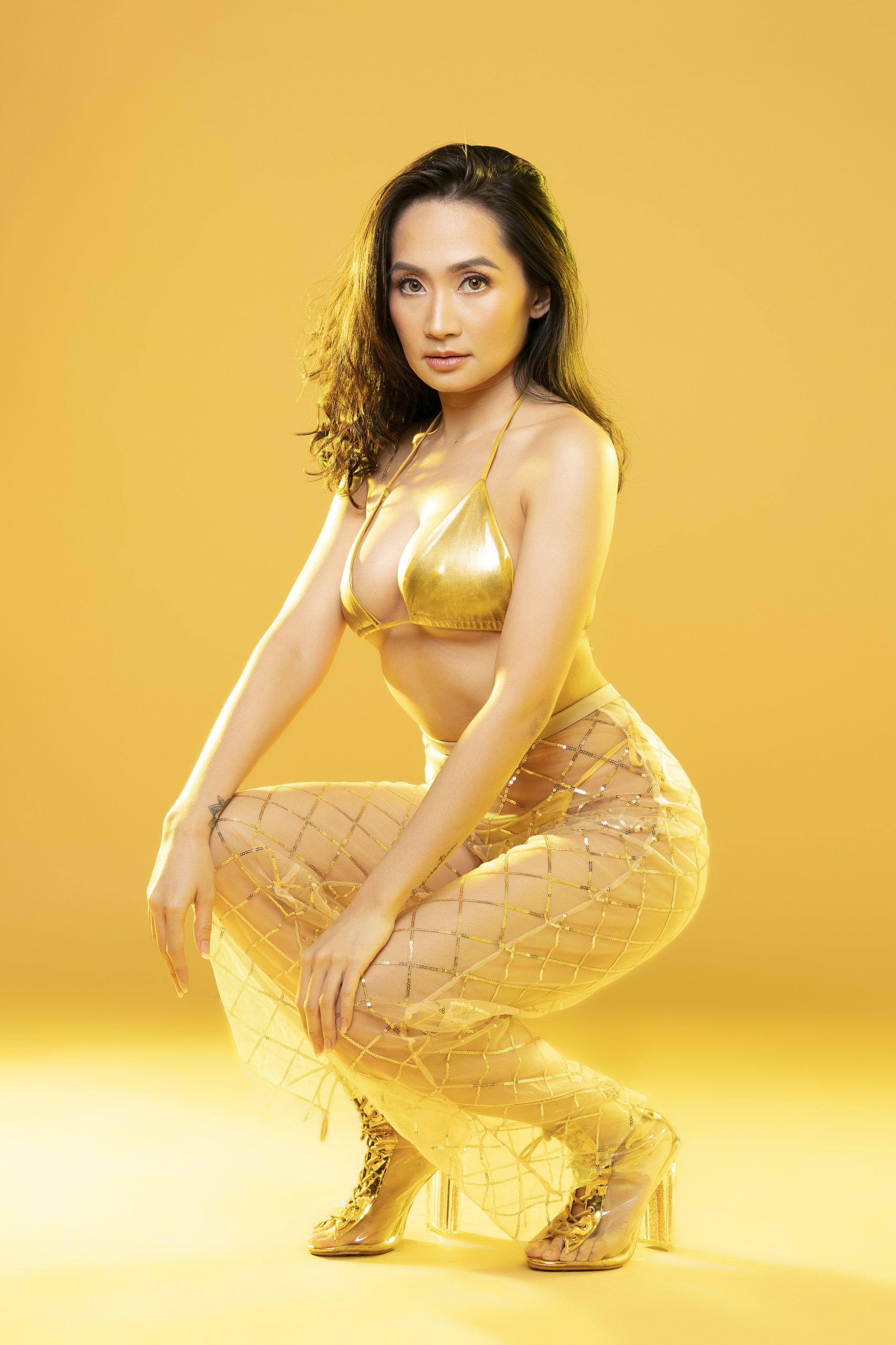 A woman with long dark hair wearing gold clothing including a metallic gold bikini top and sheer gold pants with a diamond pattern, posing against a yellow background.