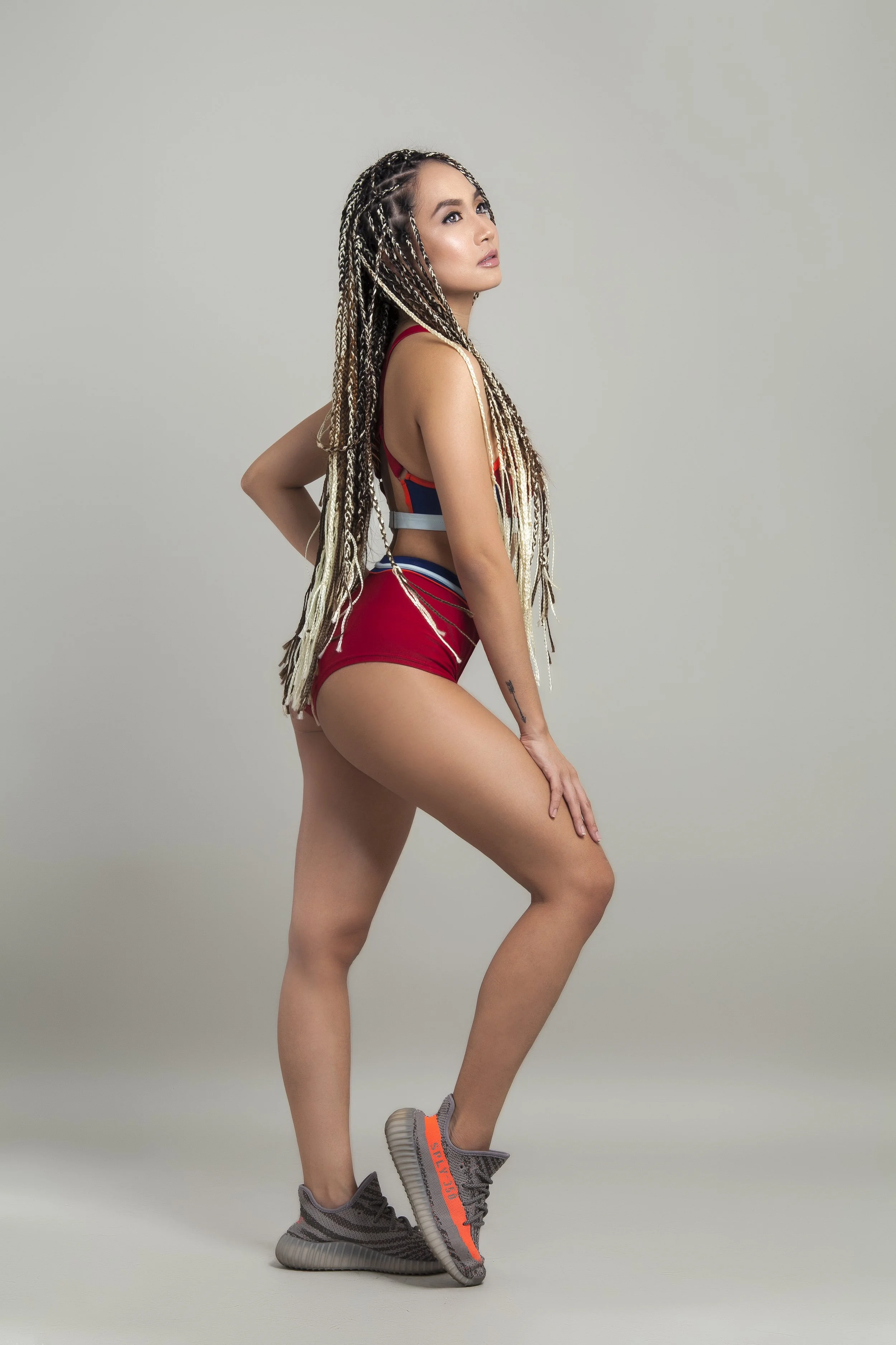 A woman with long, braided hair in various shades of blonde and brown, wearing a sporty red, blue, and gray outfit, standing in a pose with one hand behind her back and the other resting on her thigh, against a plain gray background.