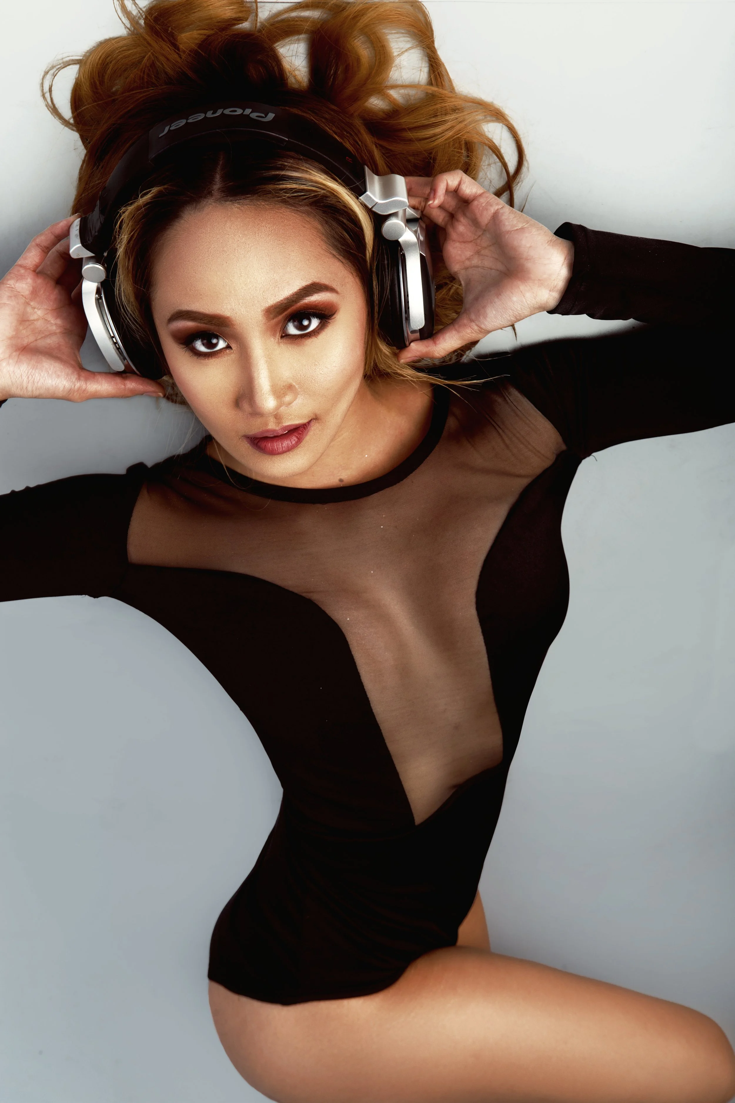 A woman with long wavy red hair, wearing a black sheer top, is lying on her back on a white surface. She is holding large headphones to her ears and looking up at the camera.