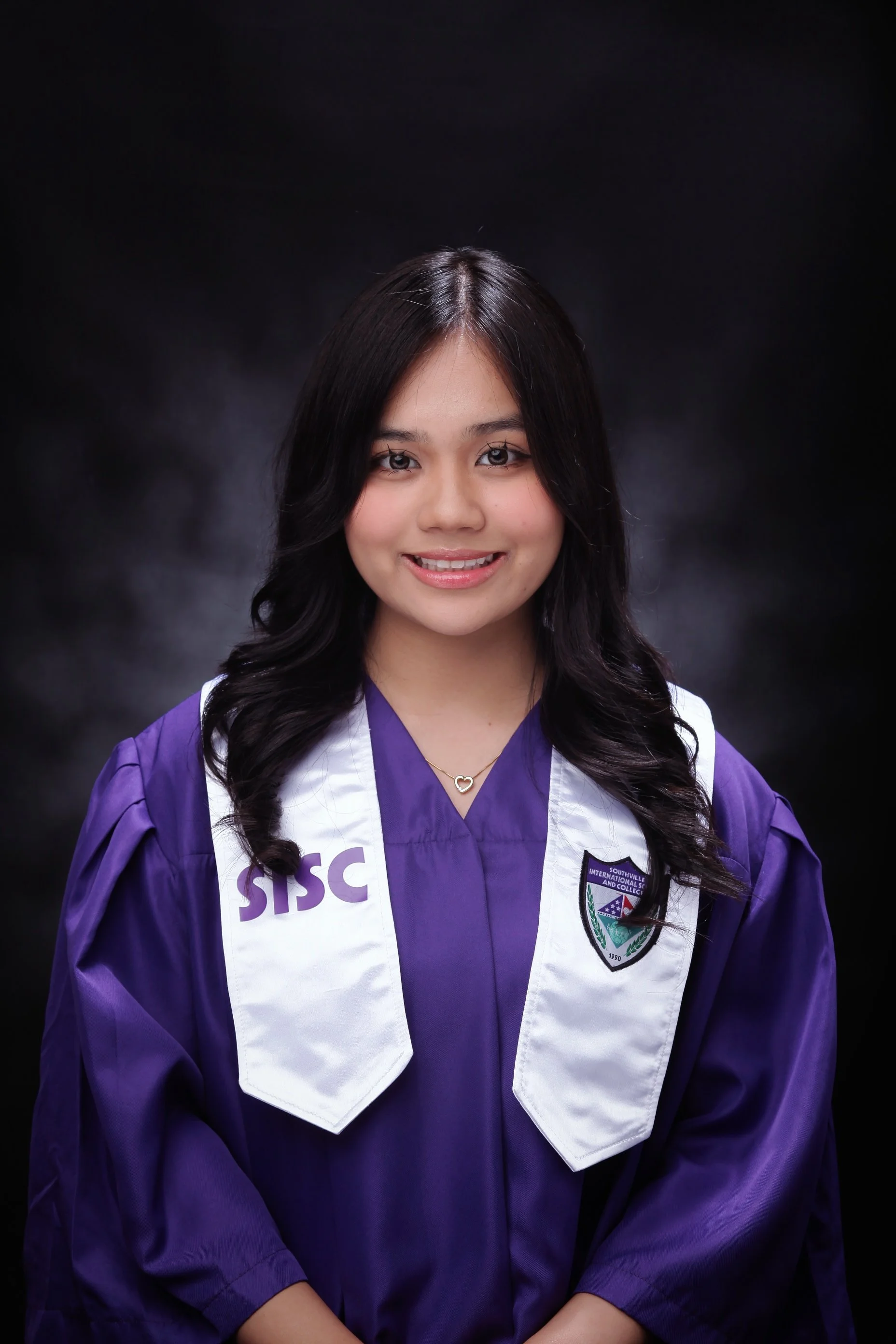 Young woman in purple graduation gown and white stole smiling, portrait against black background.