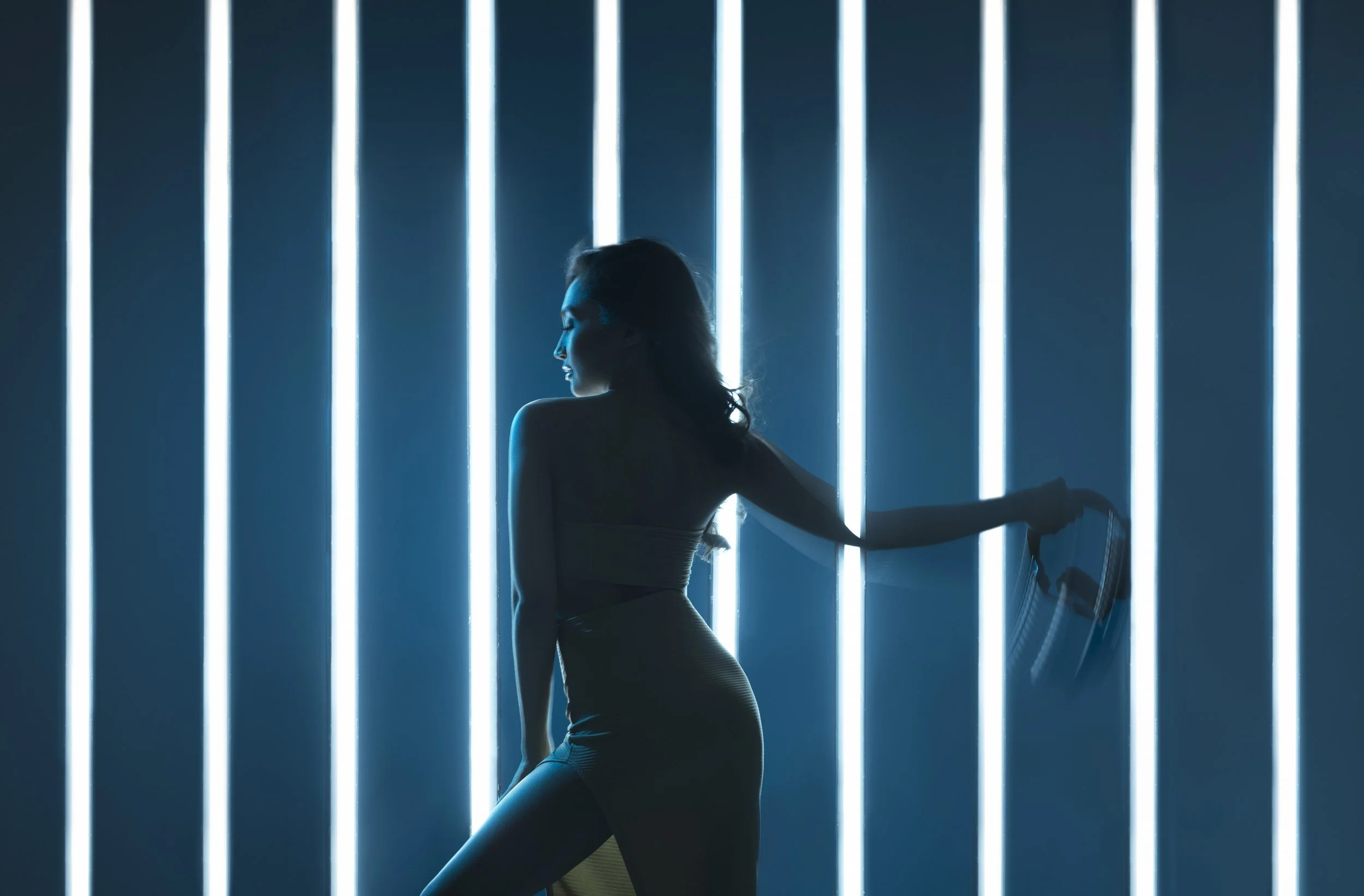 Silhouette of a woman with long hair sitting and leaning against a wall of bright blue vertical neon lights.