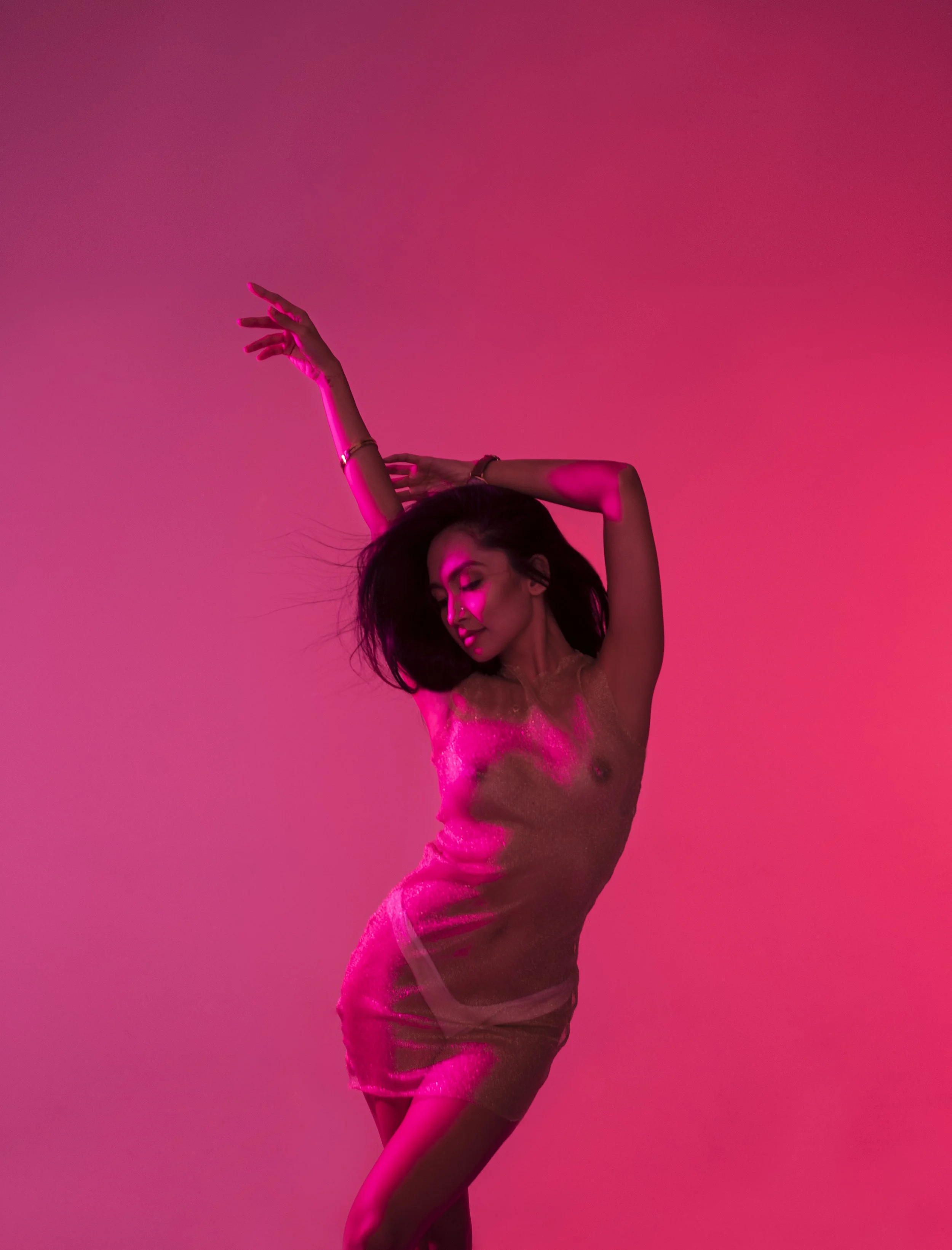 A woman dancing with one arm raised above her head, in a pink lighting setting, wearing a shimmering pink dress.