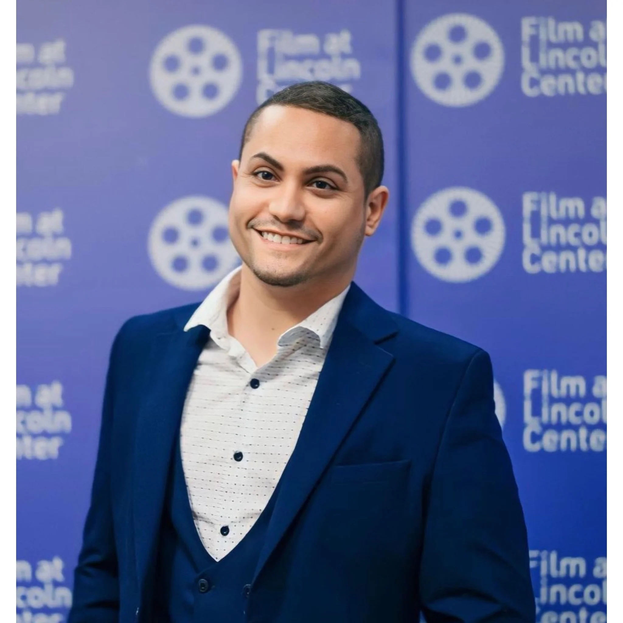 A young man dressed in a dark suit with a white shirt, smiling at a film event with a blue backdrop featuring film reel icons and the text "Film at Lincoln Center".