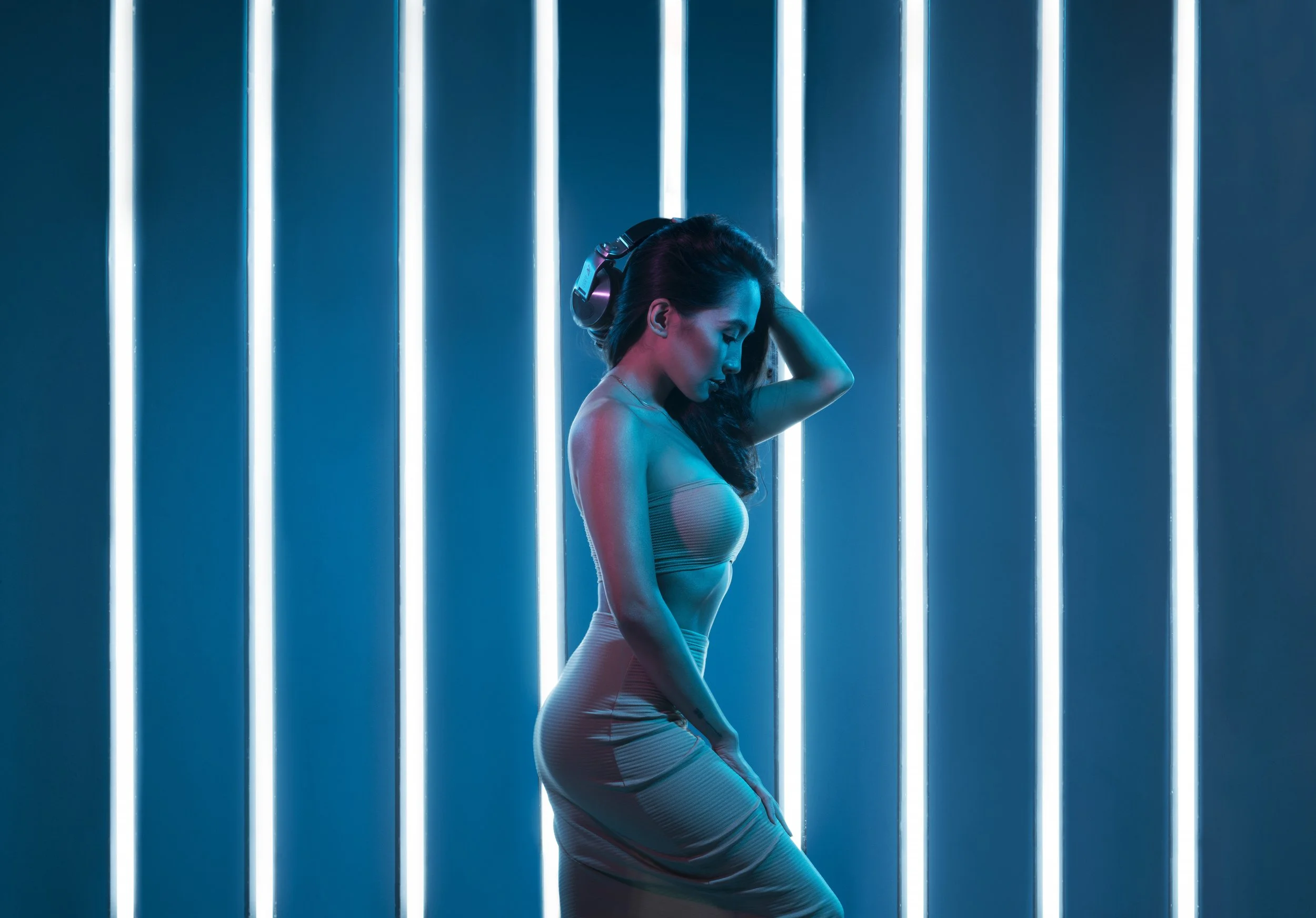 A woman with long dark hair in workout attire, wearing headphones, standing against a background of vertical neon light tubes in shades of blue and pink, with her eyes closed, touching her head gently with one hand.