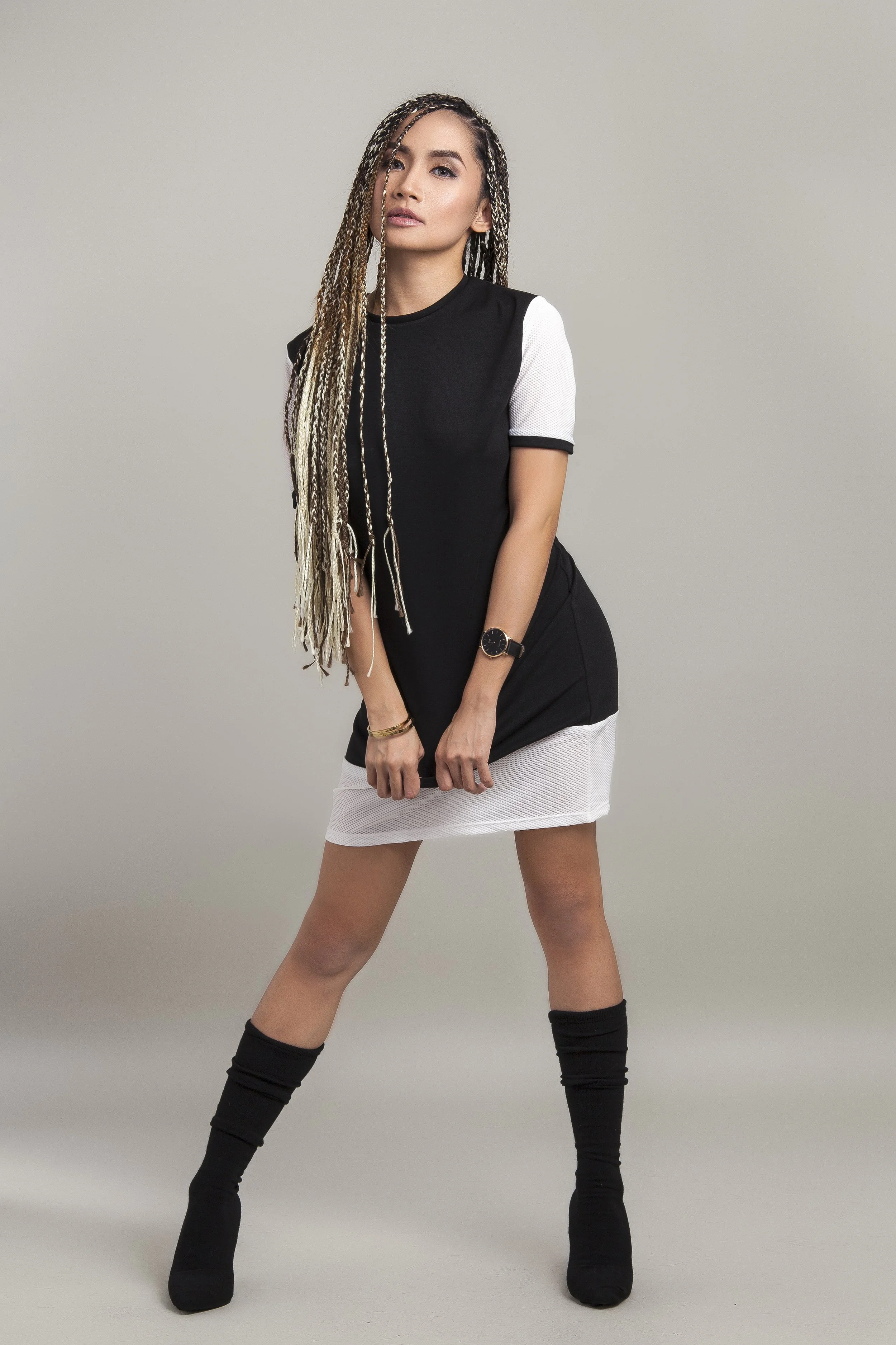 A woman with long braided hair wearing a black and white sporty dress, black knee-high socks, and black boots, standing with her legs apart against a plain light gray background.