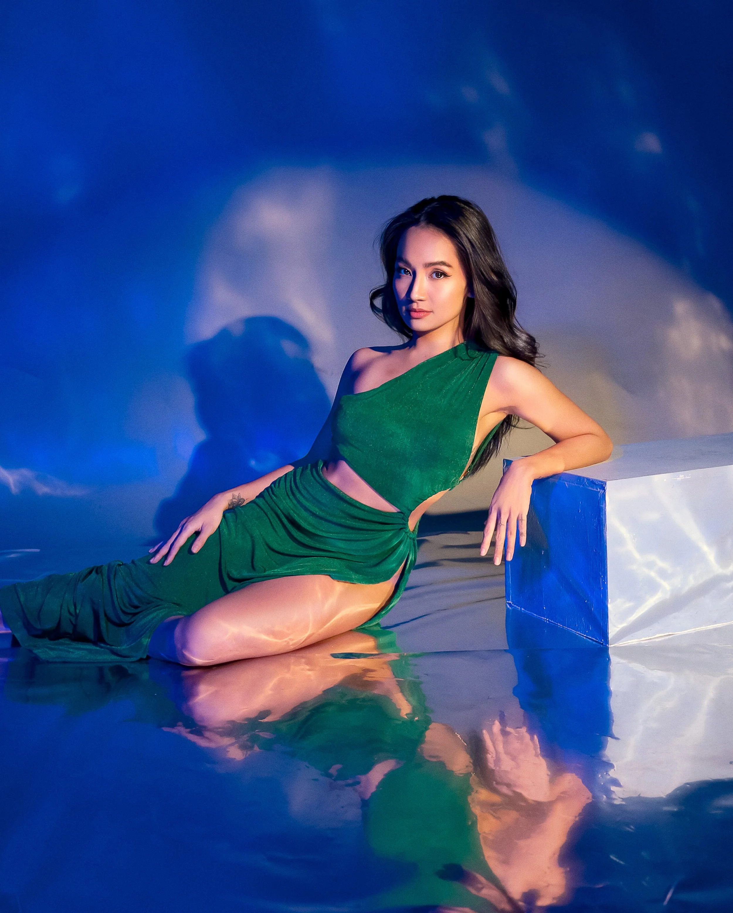 Arra Azura shot by Owen Reyes in Klickbox Studio, Pasig. She has a long dark hair and poses in front of a blue background with clouds, wearing a green dress with a cutout at the waist, sitting on the ground and leaning against a blue block.