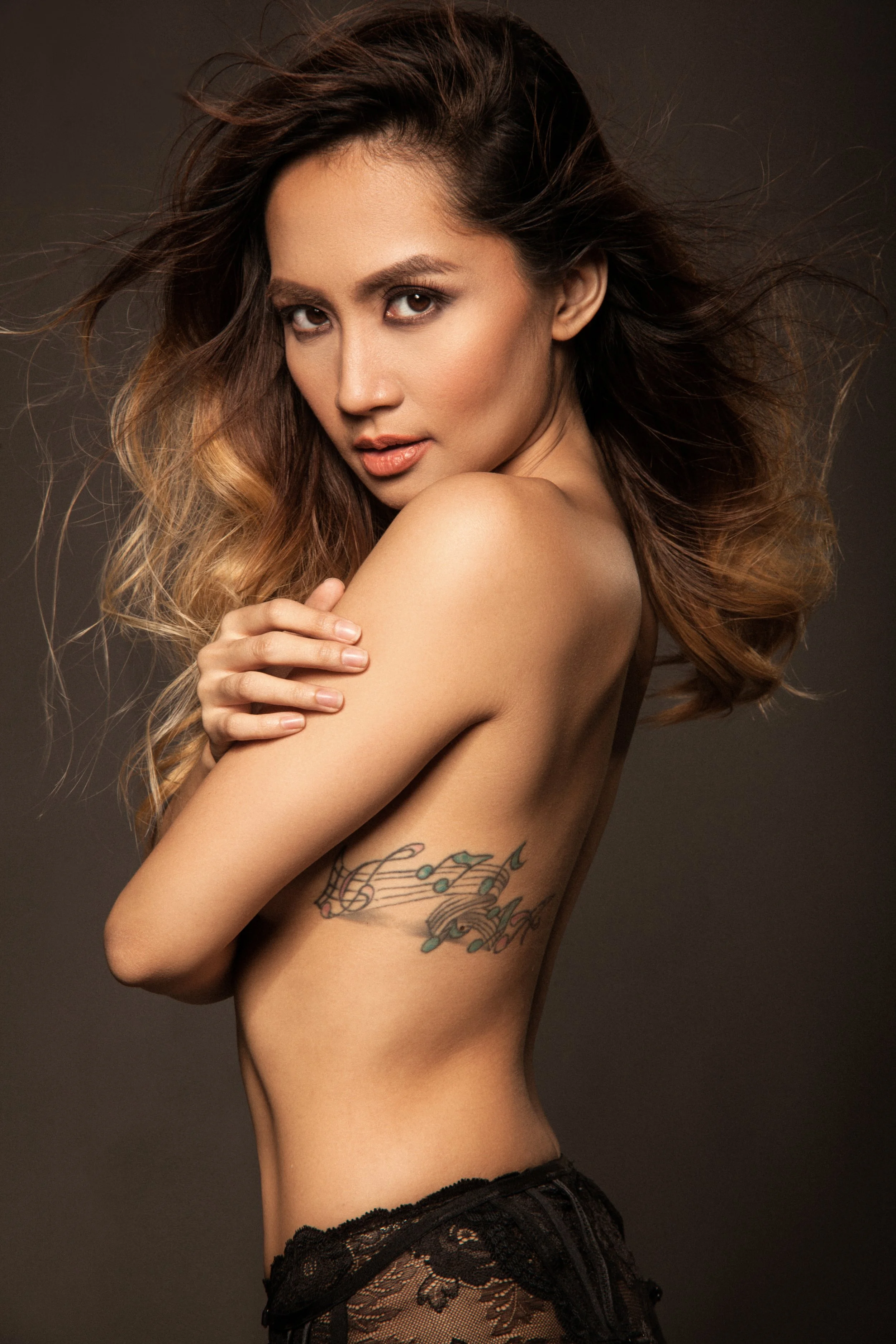 A woman with long, wavy hair and tattoos on her arm and side, posing shirtless against a dark background.