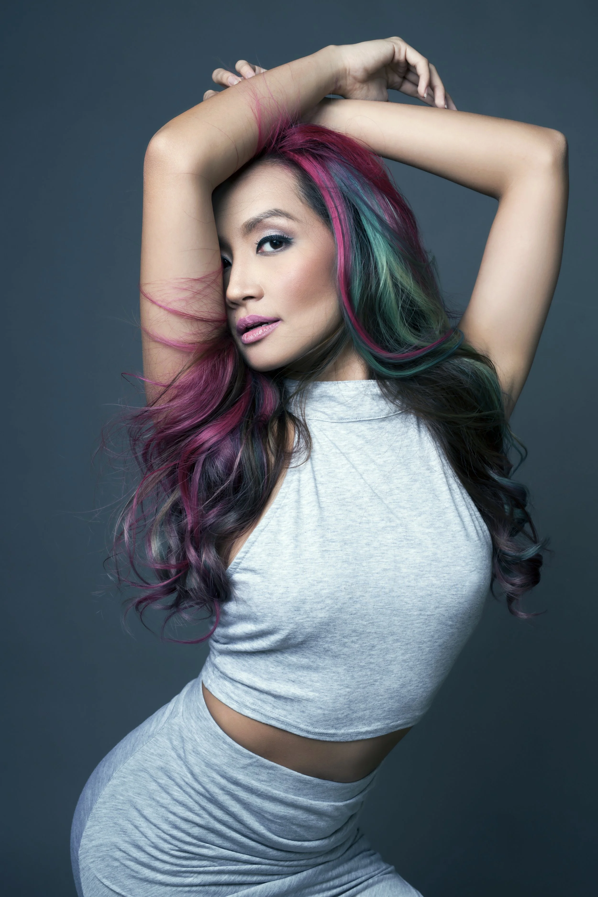 A woman with brightly colored, wavy hair in pink, blue, and green, posing with one arm raised and her face turned slightly to the side, against a solid gray background.