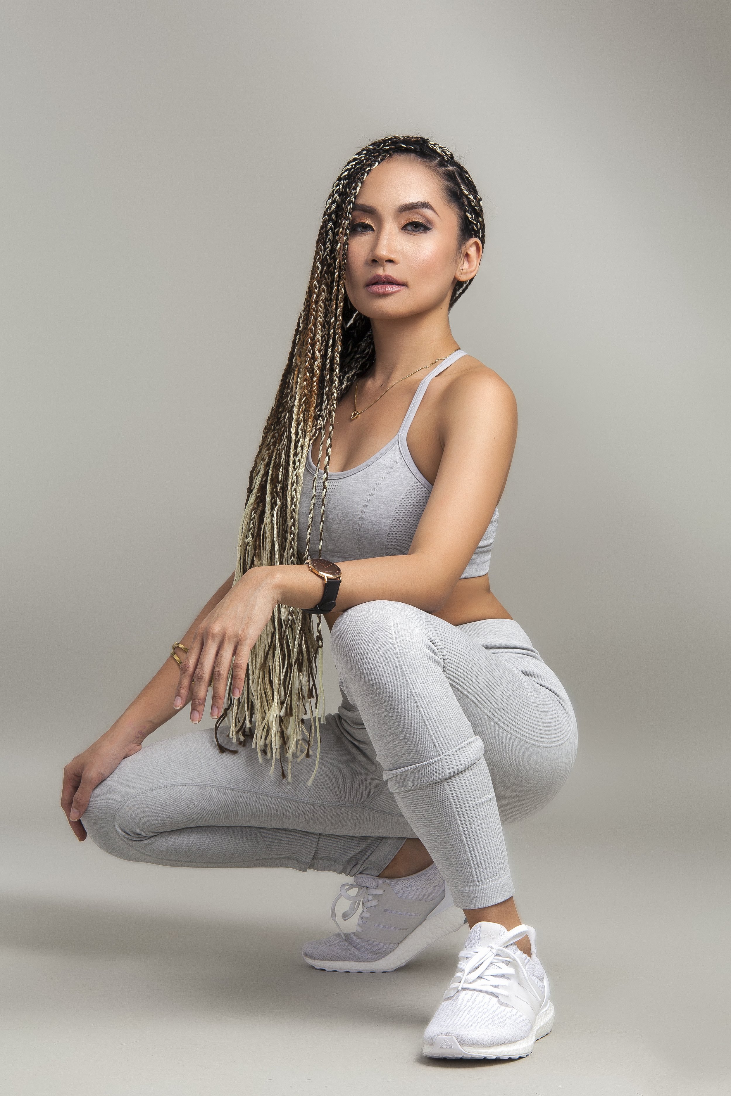 A woman wearing gray athletic wear, including a tank top, leggings, and sneakers, squatting with her right arm resting on her knee against a plain gray background.