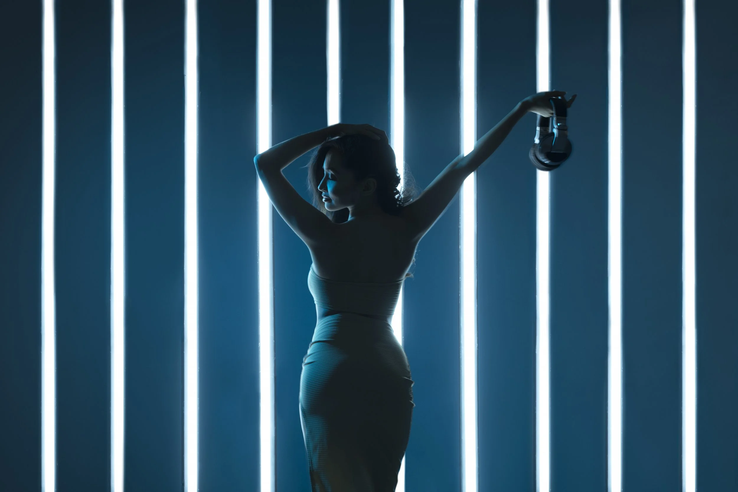 A silhouette of a woman holding a pair of headphones, standing against a backdrop of vertical blue neon light strips in a dark setting.