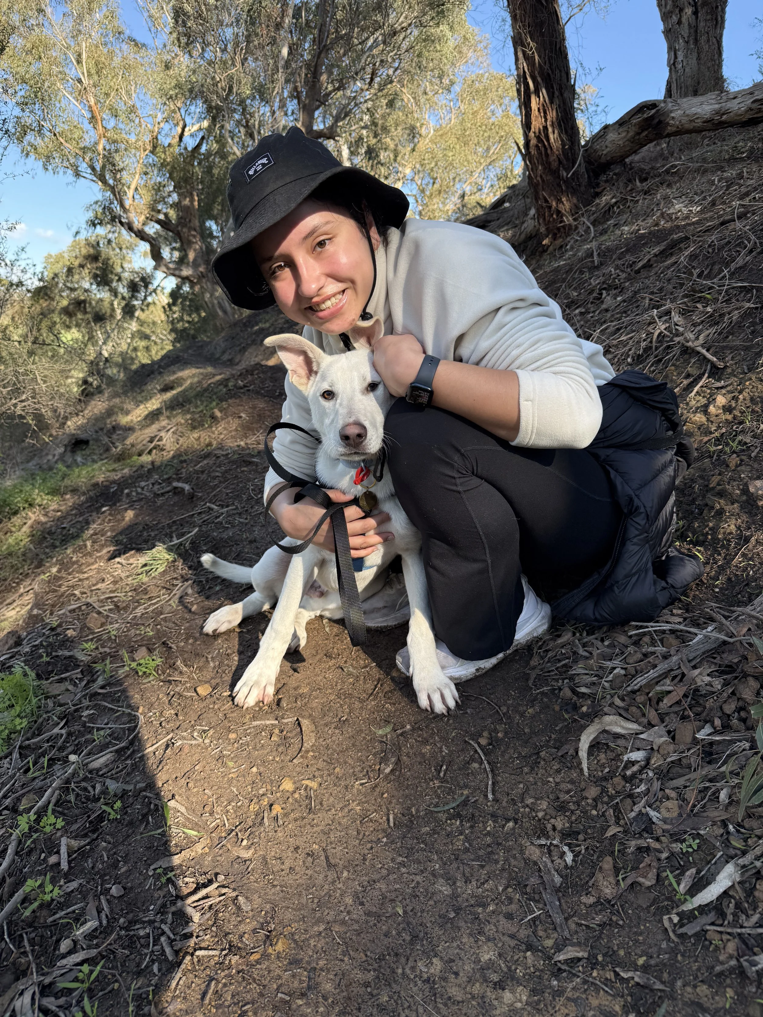 A person smiling while kneeling outdoors on a dirt trail, holding a white dog with a black collar and a red tag, surrounded by trees under a clear blue sky.