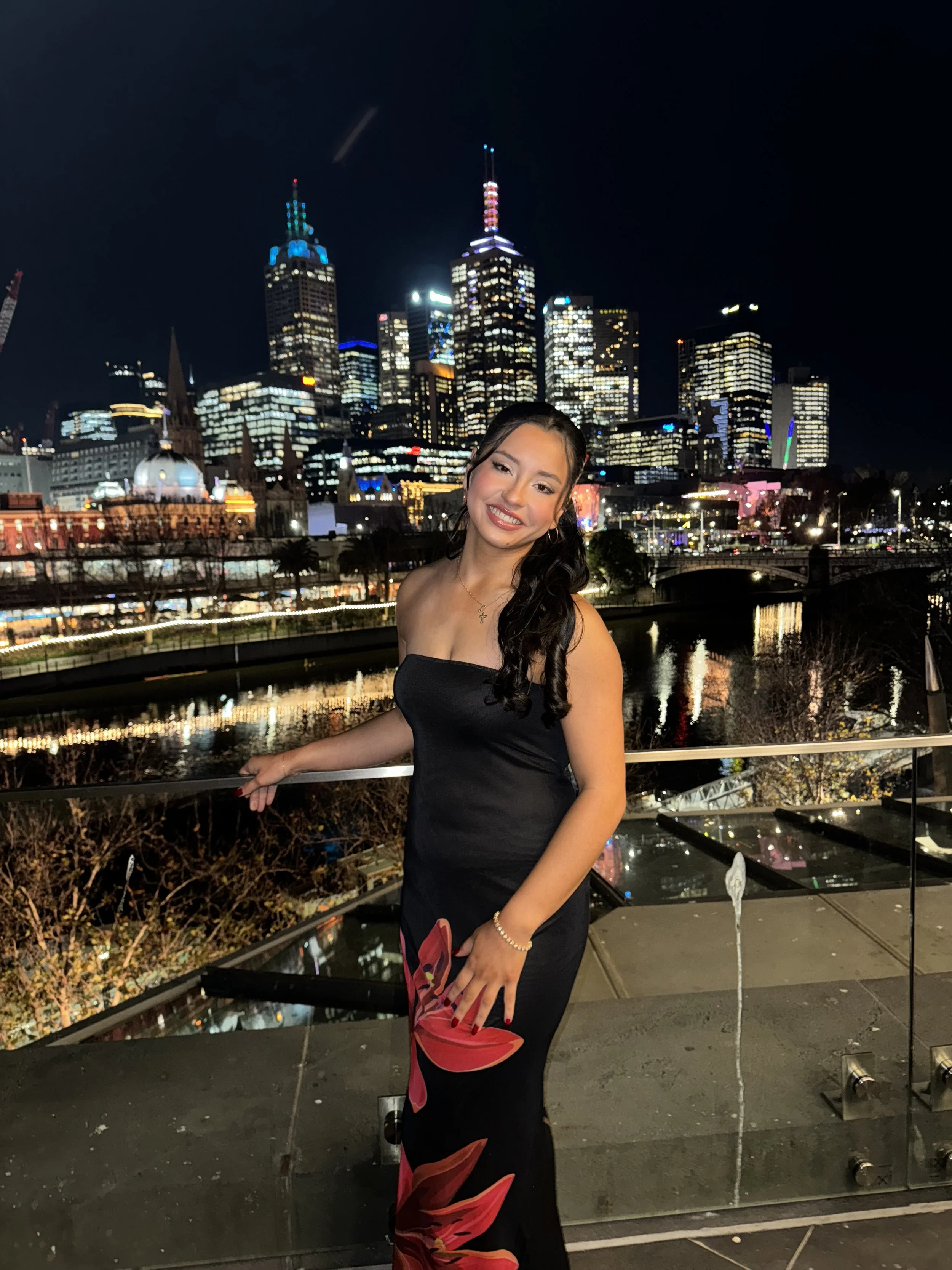 A woman in a black dress with red and orange floral patterns standing outdoors at night in front of a city skyline with illuminated skyscrapers and a river.