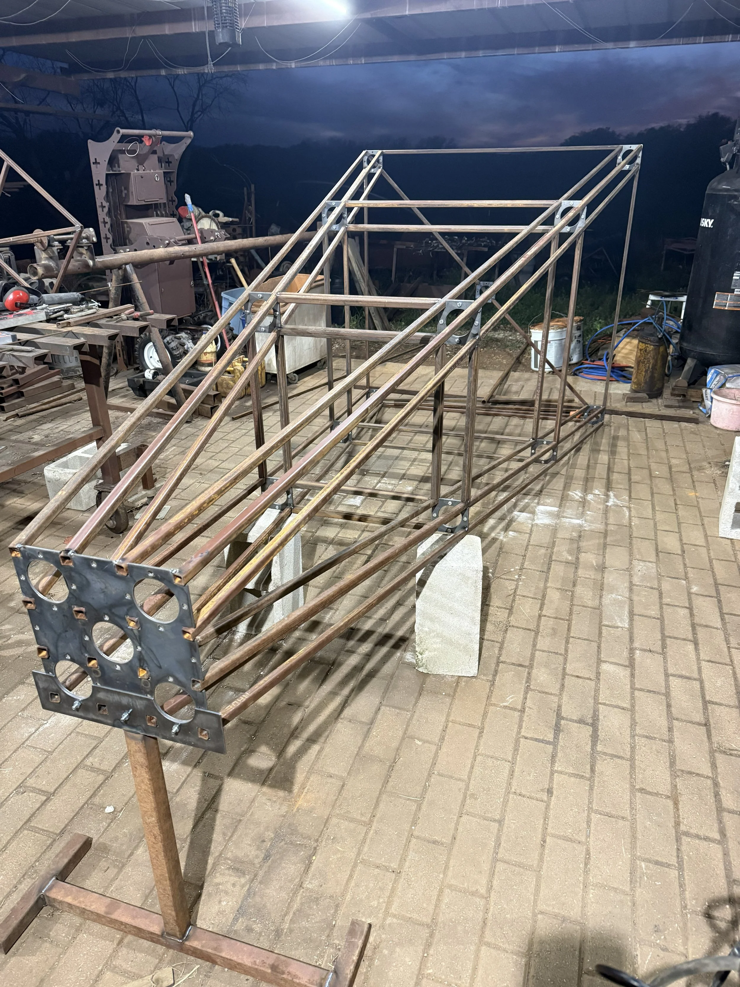A metal frame structure is being built, supported by concrete blocks, with a workshop in the background containing tools, equipment, and supplies.